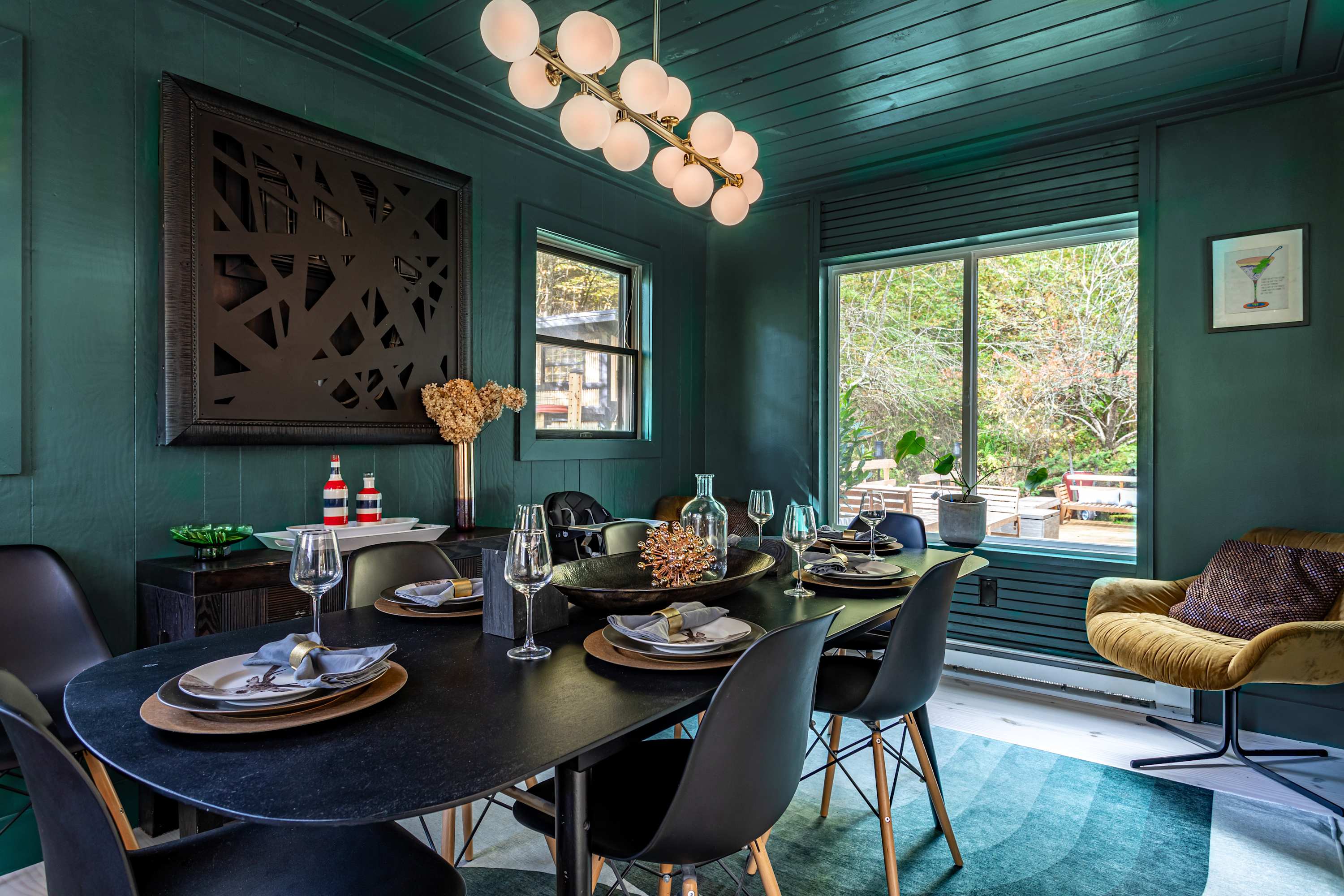 Dining room with dark teal walls and ceiling, a black oval dining table with matching chairs, and a modern globe chandelier, featuring a large abstract black artwork and a window overlooking the outdoors.