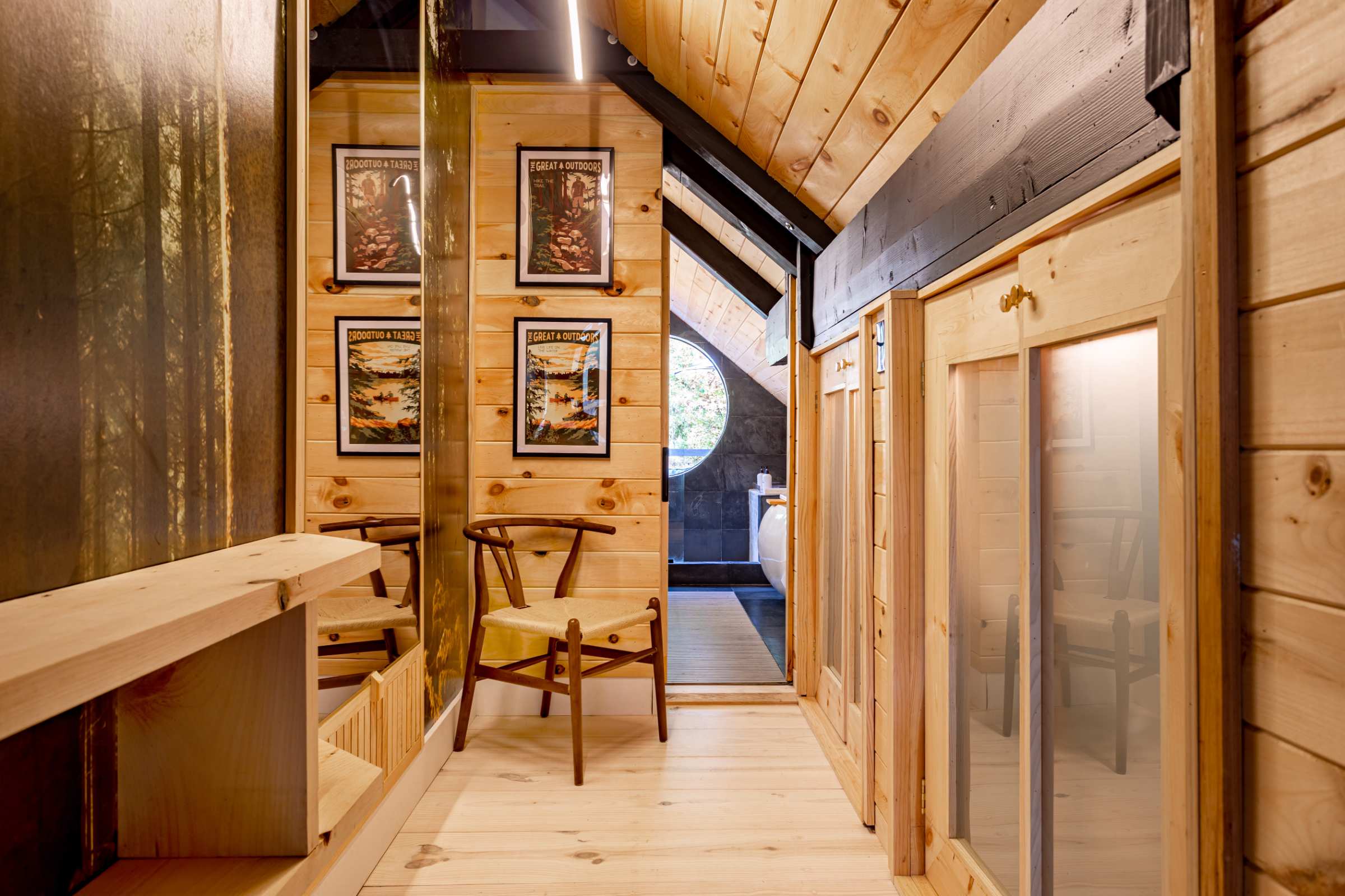 A narrow hallway with light wood paneling on the walls and ceiling, featuring framed artwork, a full-length mirror, and leading towards a bathroom with a round window.