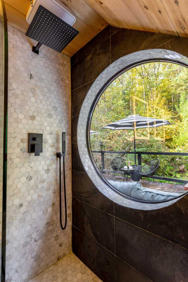 Modern shower with hexagonal white and grey mosaic tiles, a black rainfall showerhead, and a black handheld shower. A large circular window offers a view of an outdoor deck with a striped umbrella and lush trees.