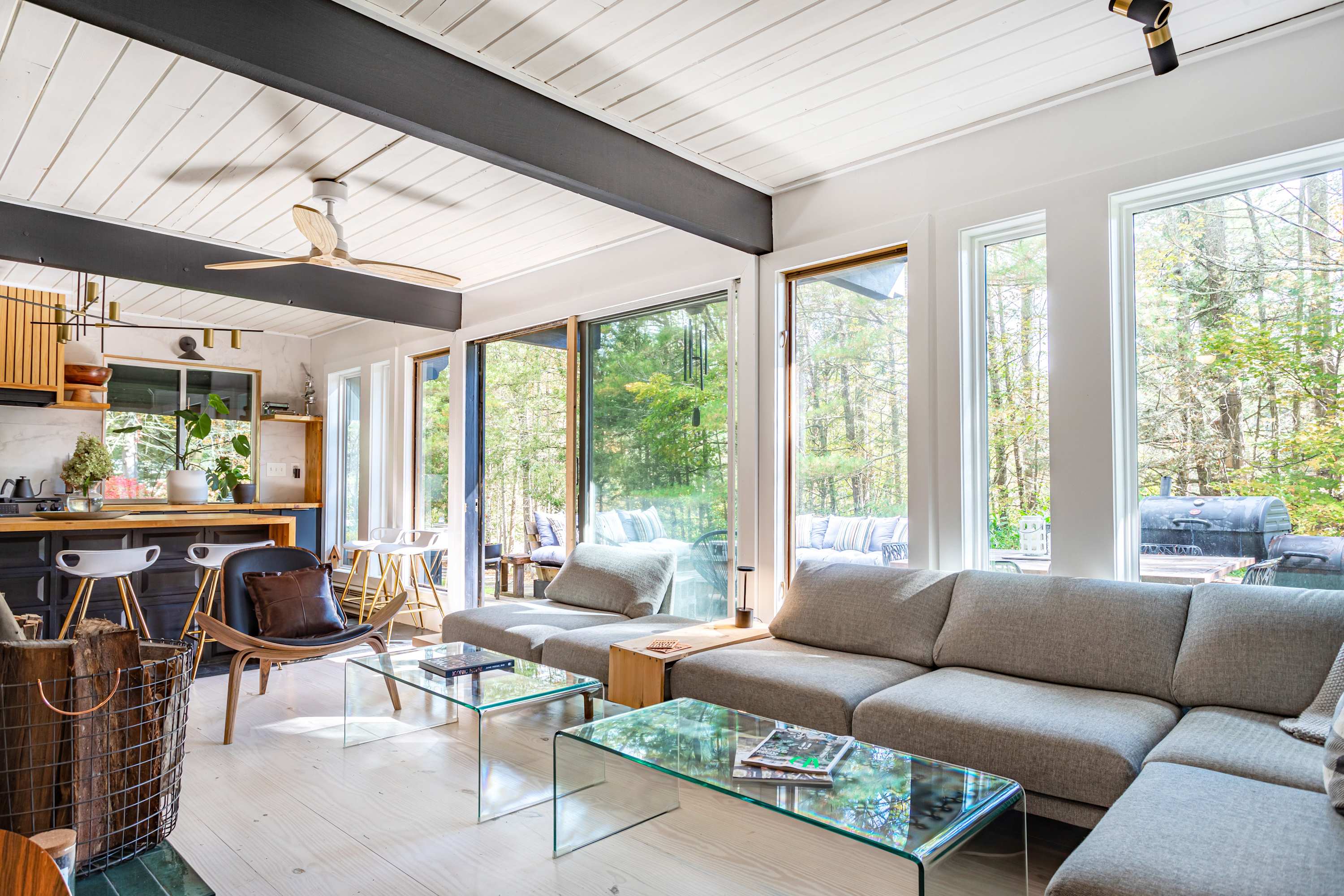 Open concept living space featuring a gray sectional sofa, glass coffee tables, and large windows overlooking a wooded area, with a glimpse of the kitchen island and a wooden ceiling fan.