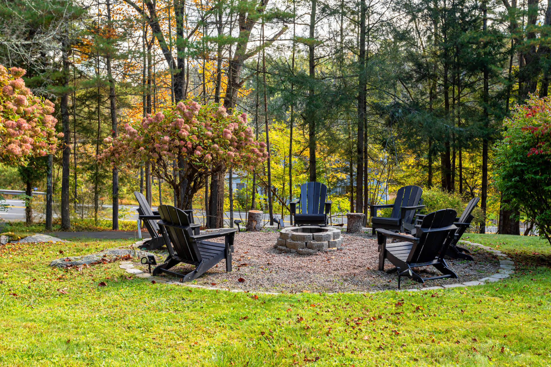 Circular outdoor fire pit area surrounded by black Adirondack chairs on a gravel base, set within a grassy yard with mature trees displaying autumn colors.