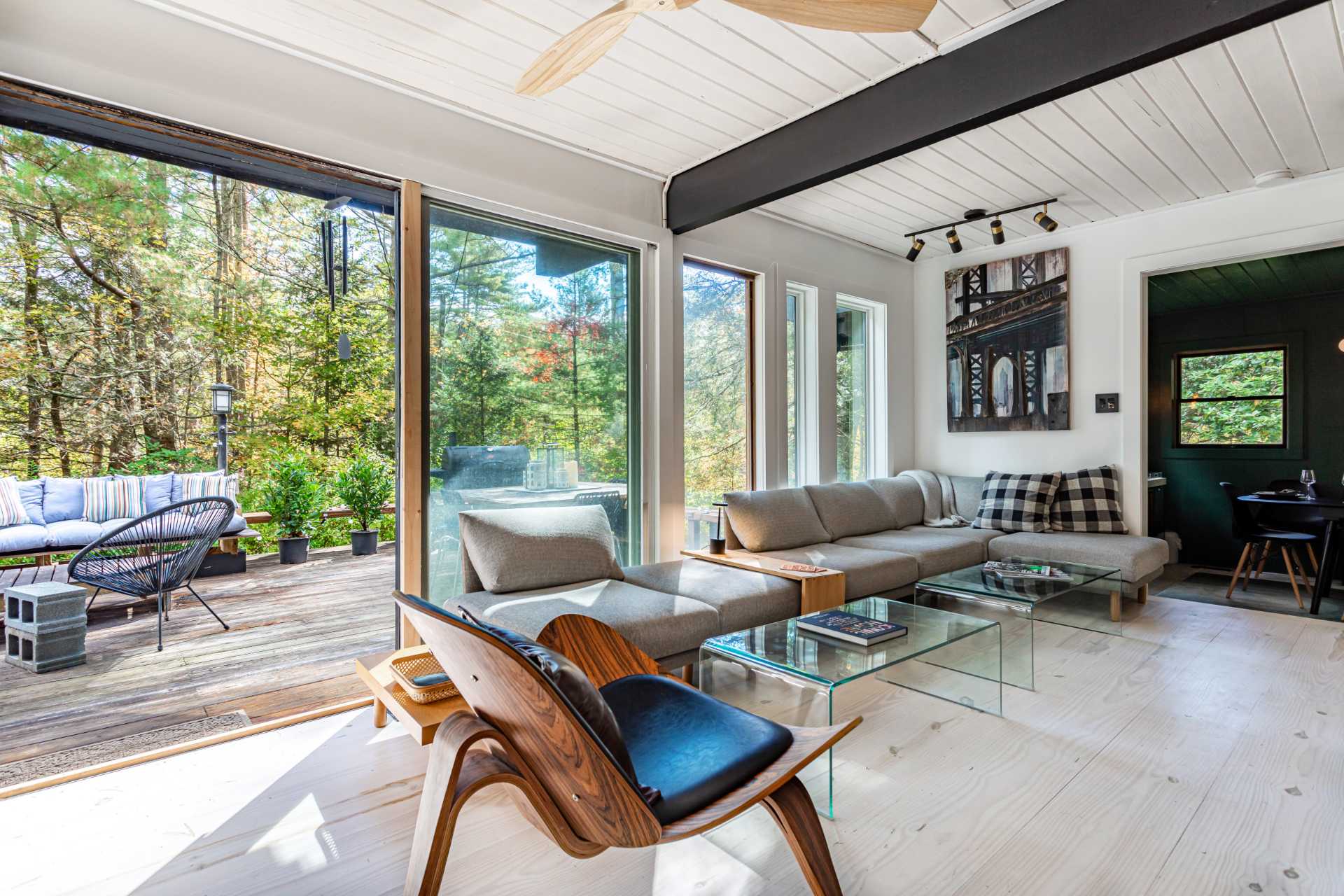 Bright living room with white paneled ceilings, a large L-shaped gray sectional sofa, glass coffee tables, and a modern wooden chair, all facing large glass doors and windows that open to a wooden deck and forest views.
