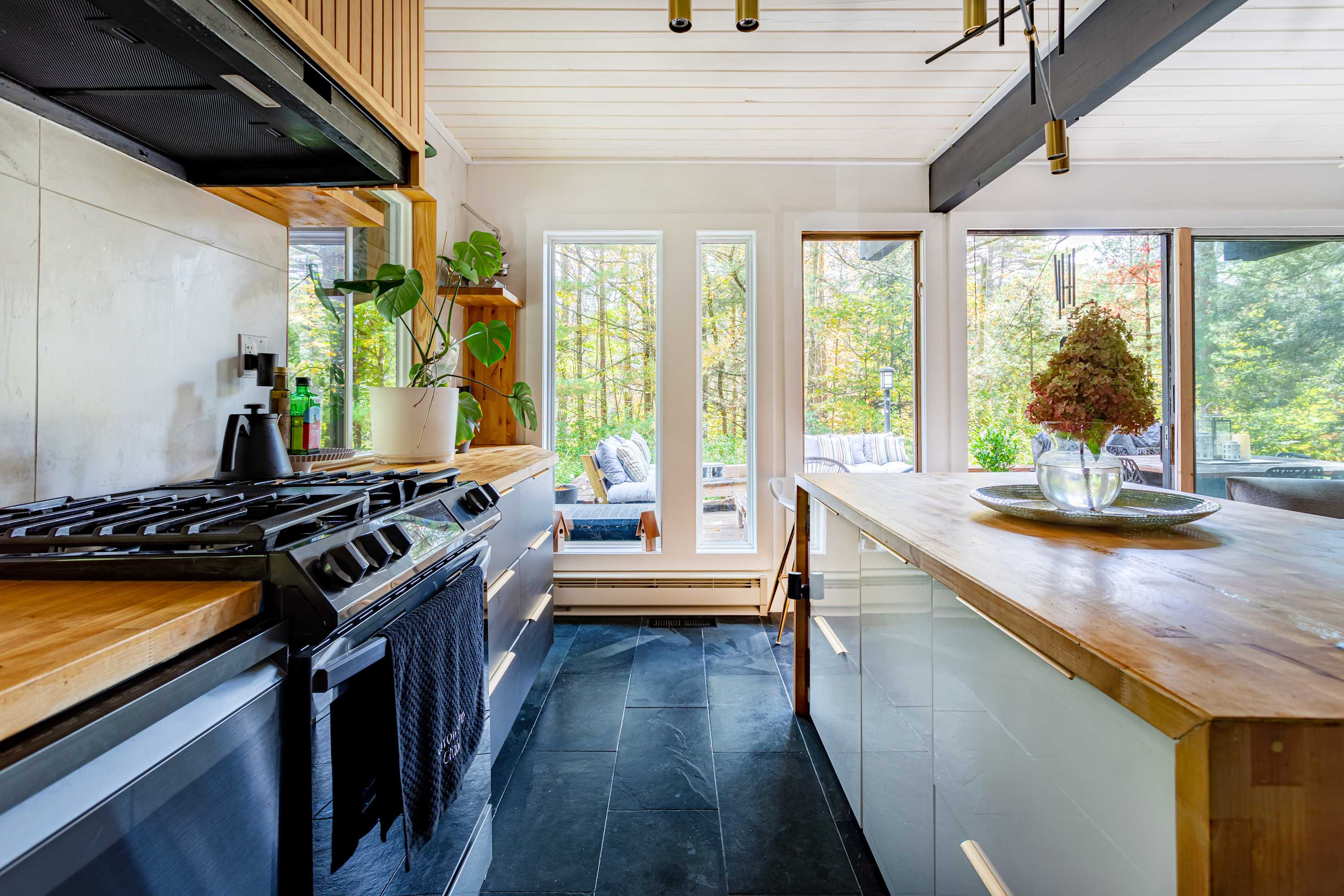 Close-up of the kitchen area featuring a black gas range, dark slate floor tiles, wooden countertops, and large windows looking out to the deck and surrounding trees.