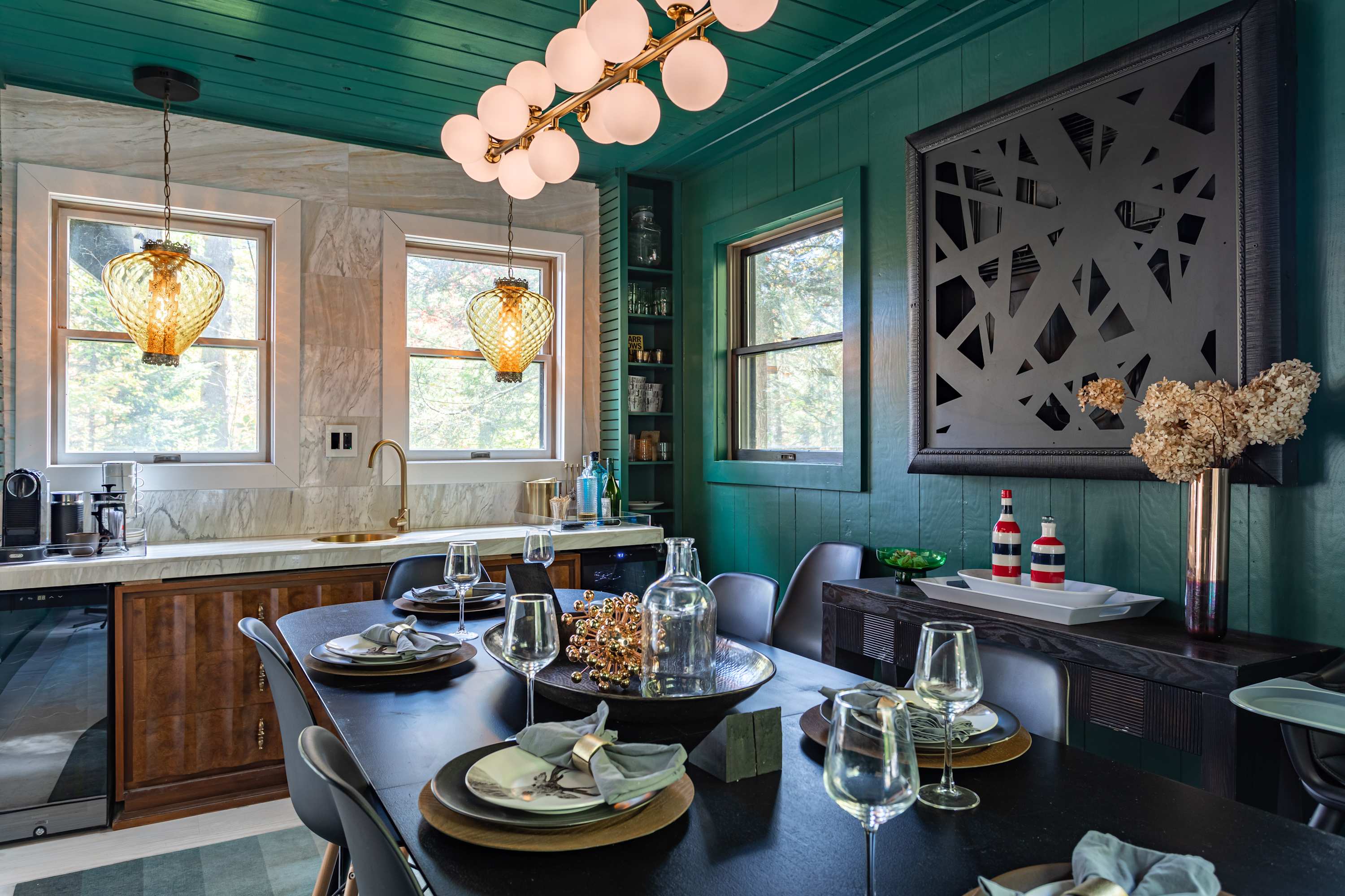 Elegant dining room with dark teal walls, a black dining table set for guests, and a wet bar area with marble backsplash and two decorative pendant lights, all under a modern globe chandelier.