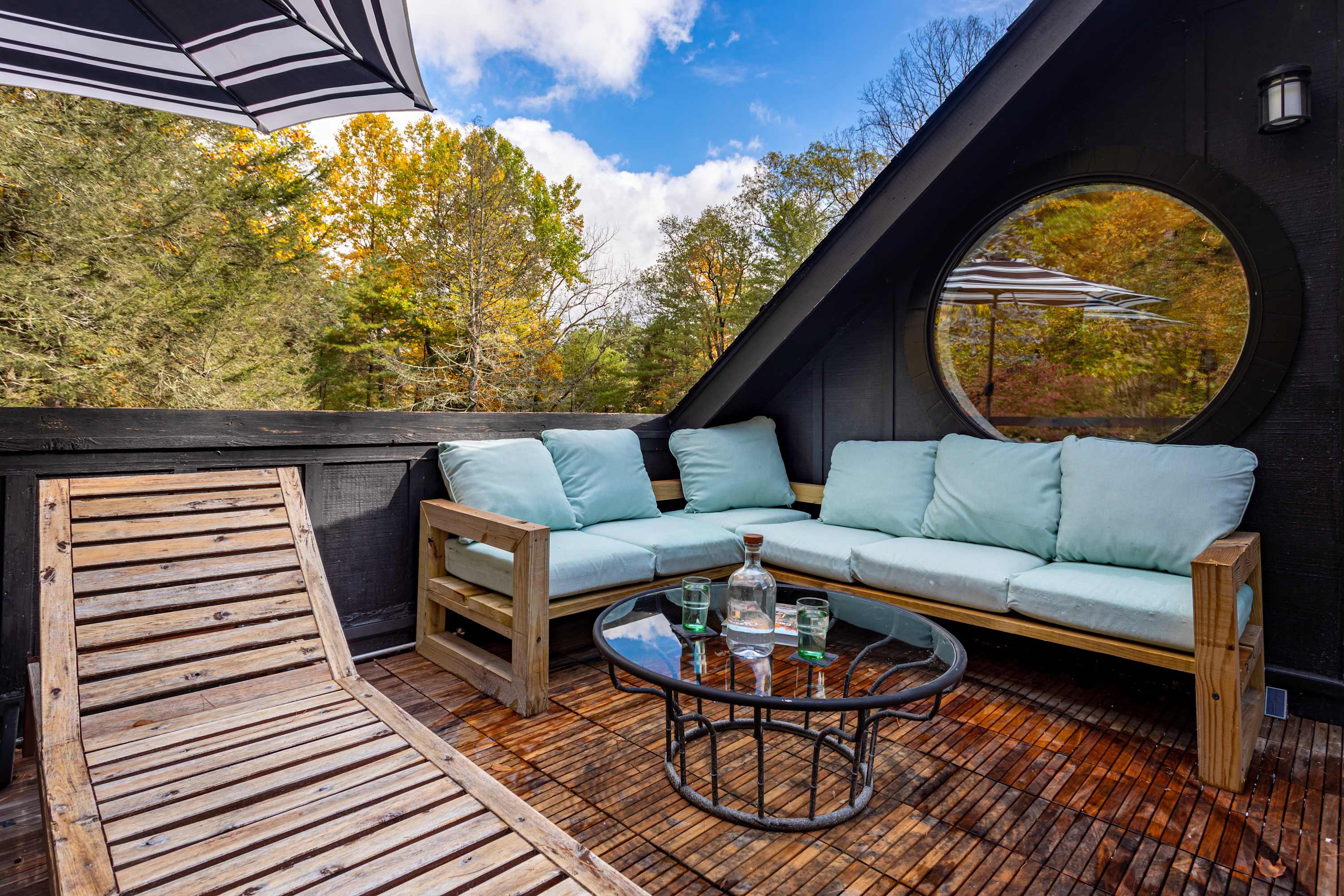 Spacious outdoor deck with a light blue cushioned wooden sectional sofa and a glass-top coffee table, set against a backdrop of autumn trees. A black and white striped umbrella provides shade, and a circular window reflects the outdoor scene.