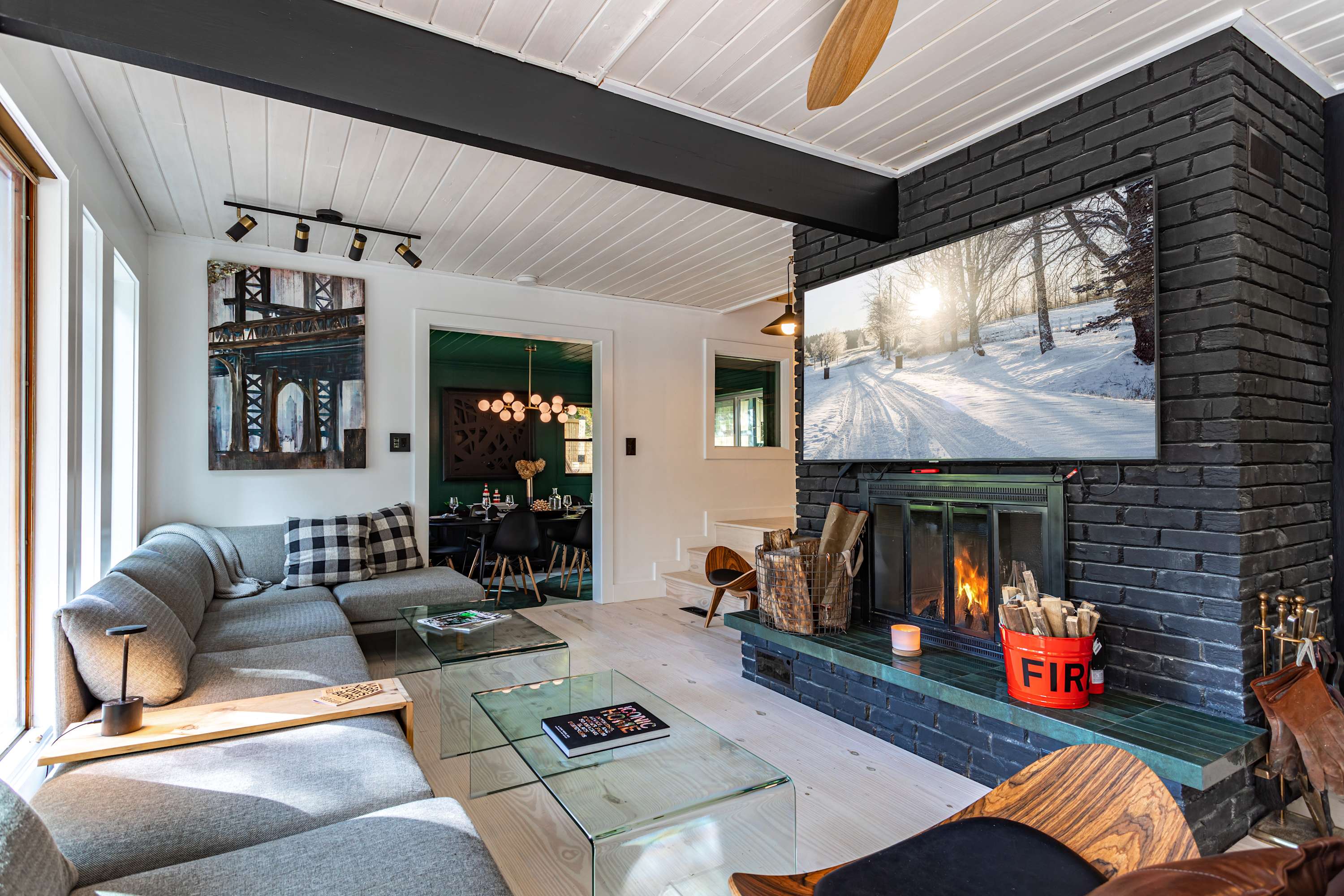 Cozy living room with a large grey sectional sofa, glass coffee tables, and a black brick fireplace with a mounted TV, featuring white planked ceilings and a view into a green dining room.