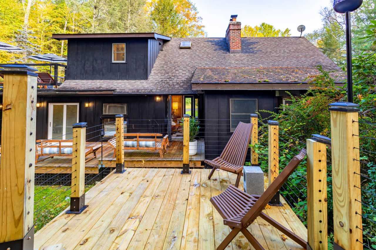 Rear view of a dark-colored house with multiple wooden decks and cable railings, surrounded by lush green and autumn-colored trees. Various outdoor seating options are visible on the decks.