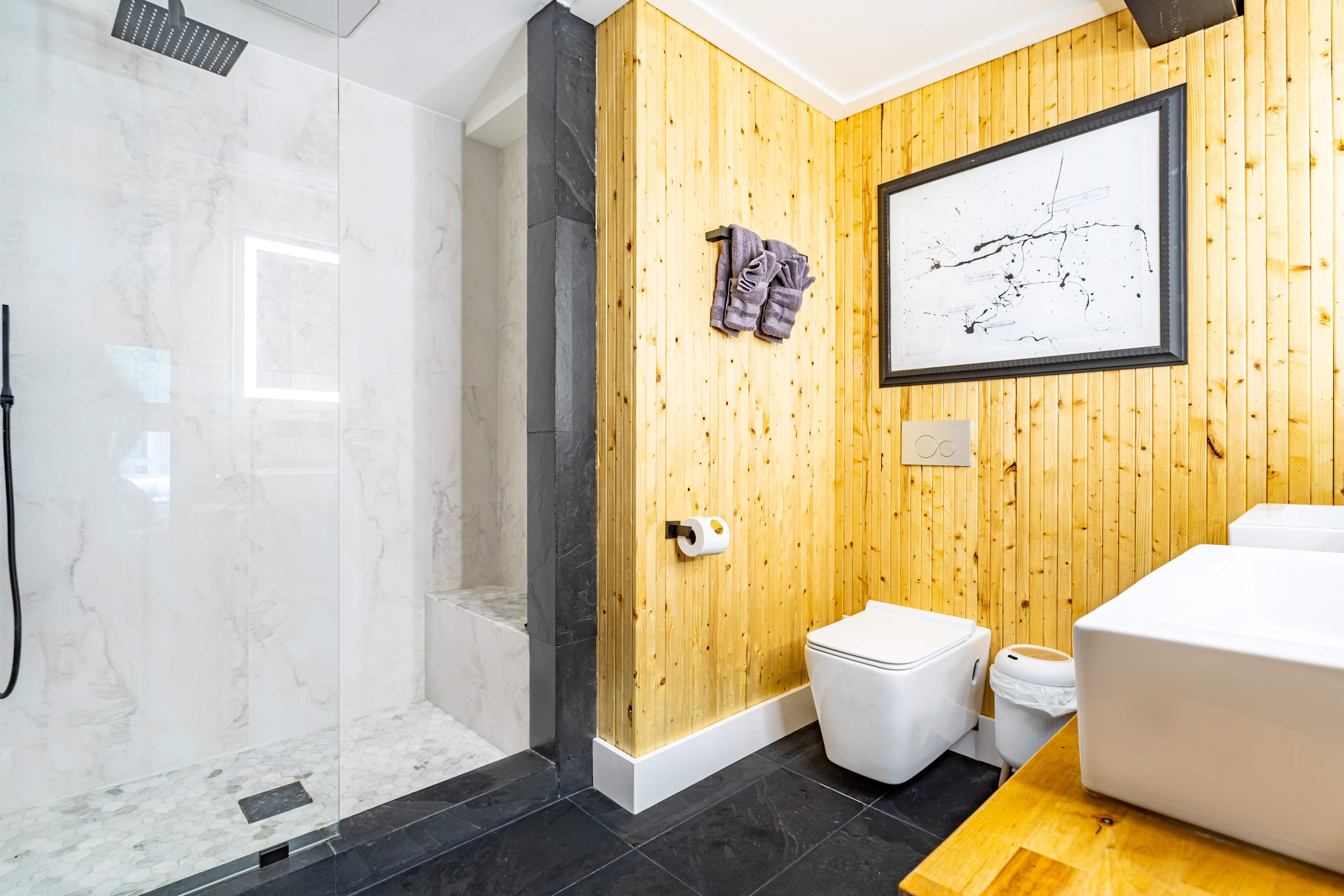 A modern bathroom with a glass-enclosed shower featuring white marble tiles and a rain showerhead, alongside a wall-mounted toilet and a wooden slatted wall.