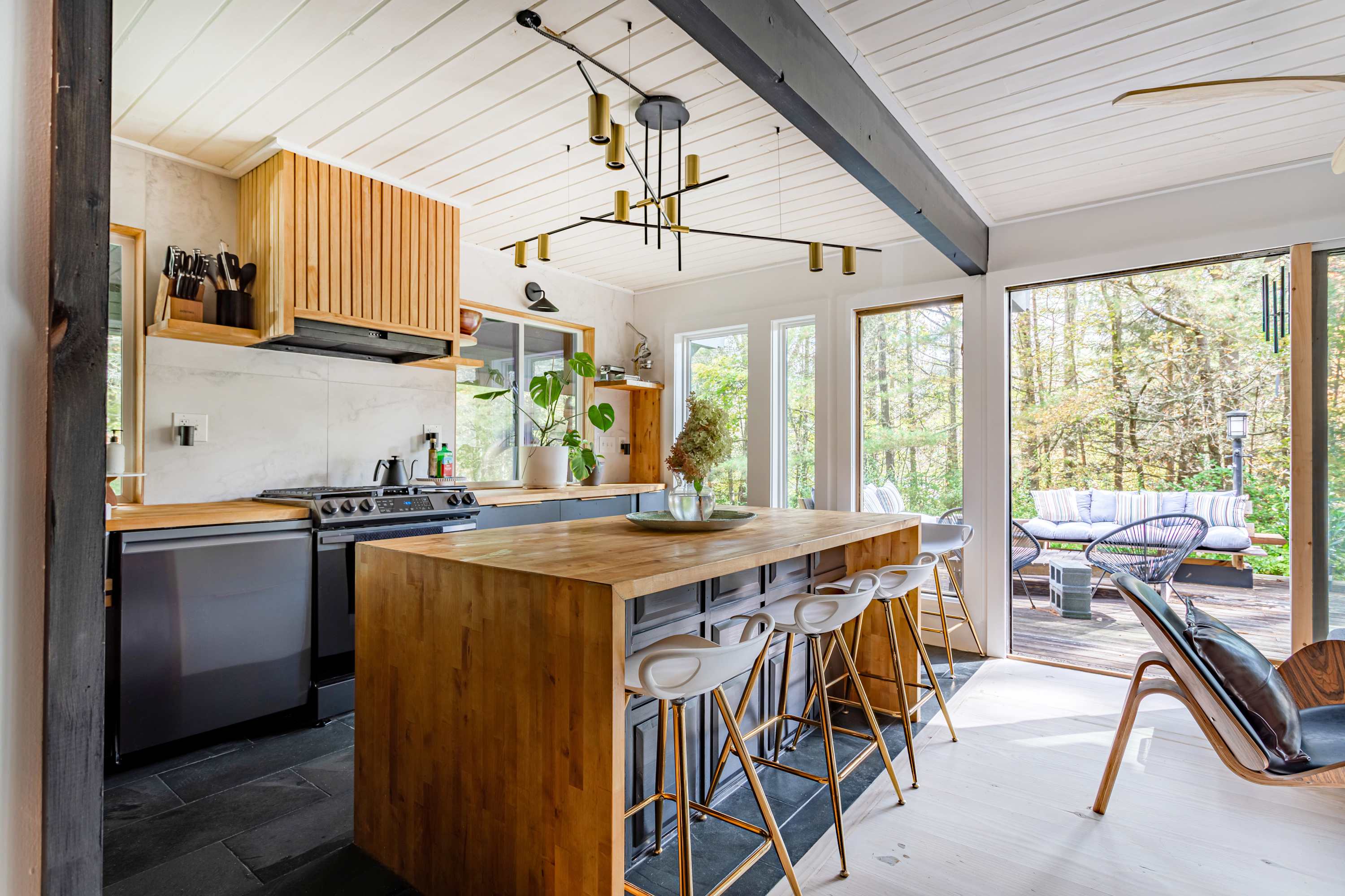 Modern kitchen with a large wooden island featuring white bar stools, black base cabinets, a gas range, and a stylish gold chandelier, all set against white paneled ceilings and light hardwood floors.