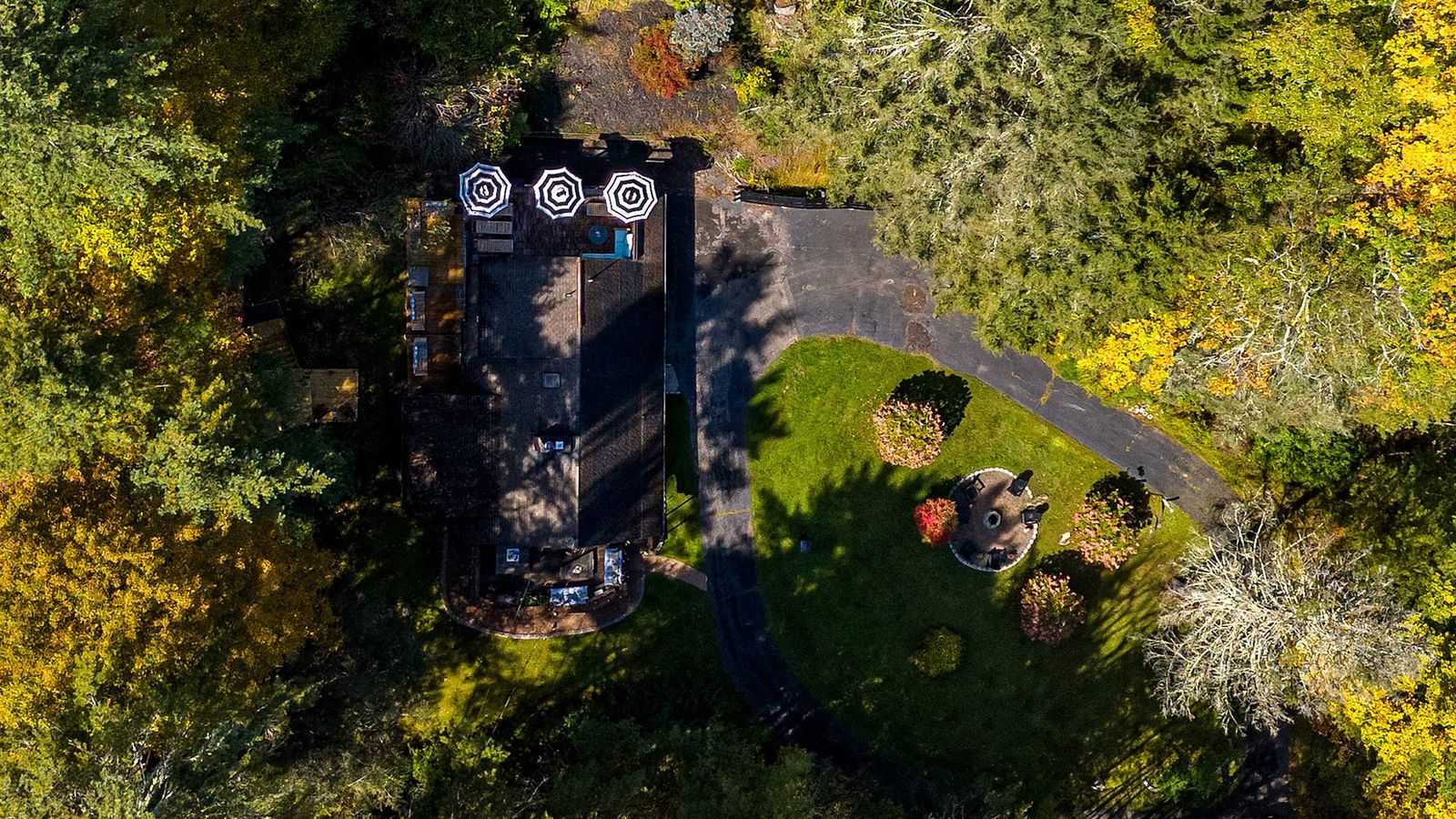 Aerial view of the property showing the dark-roofed house nestled among vibrant autumn trees, with a driveway, a fire pit area, and multiple outdoor decks with striped umbrellas.