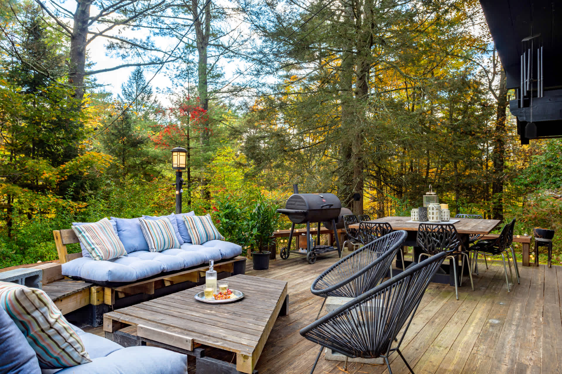 Spacious wooden deck with a large outdoor dining table and chairs, a BBQ grill, and a lounge area with a light blue cushioned sofa. The deck is surrounded by a dense forest with vibrant autumn colors.