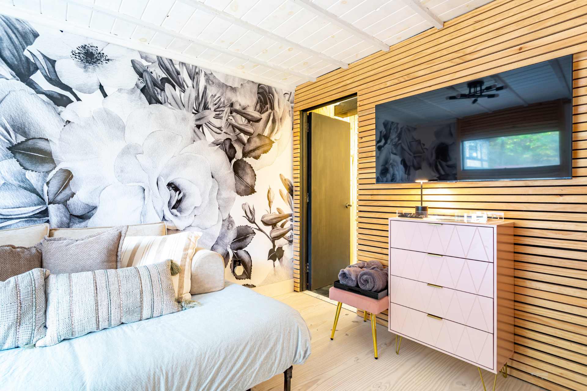 A cozy bedroom featuring a large floral mural wallpaper in black and white, a light wood slatted wall with a mounted TV, and a pink dresser with gold legs. A daybed with throw pillows is visible on the left.