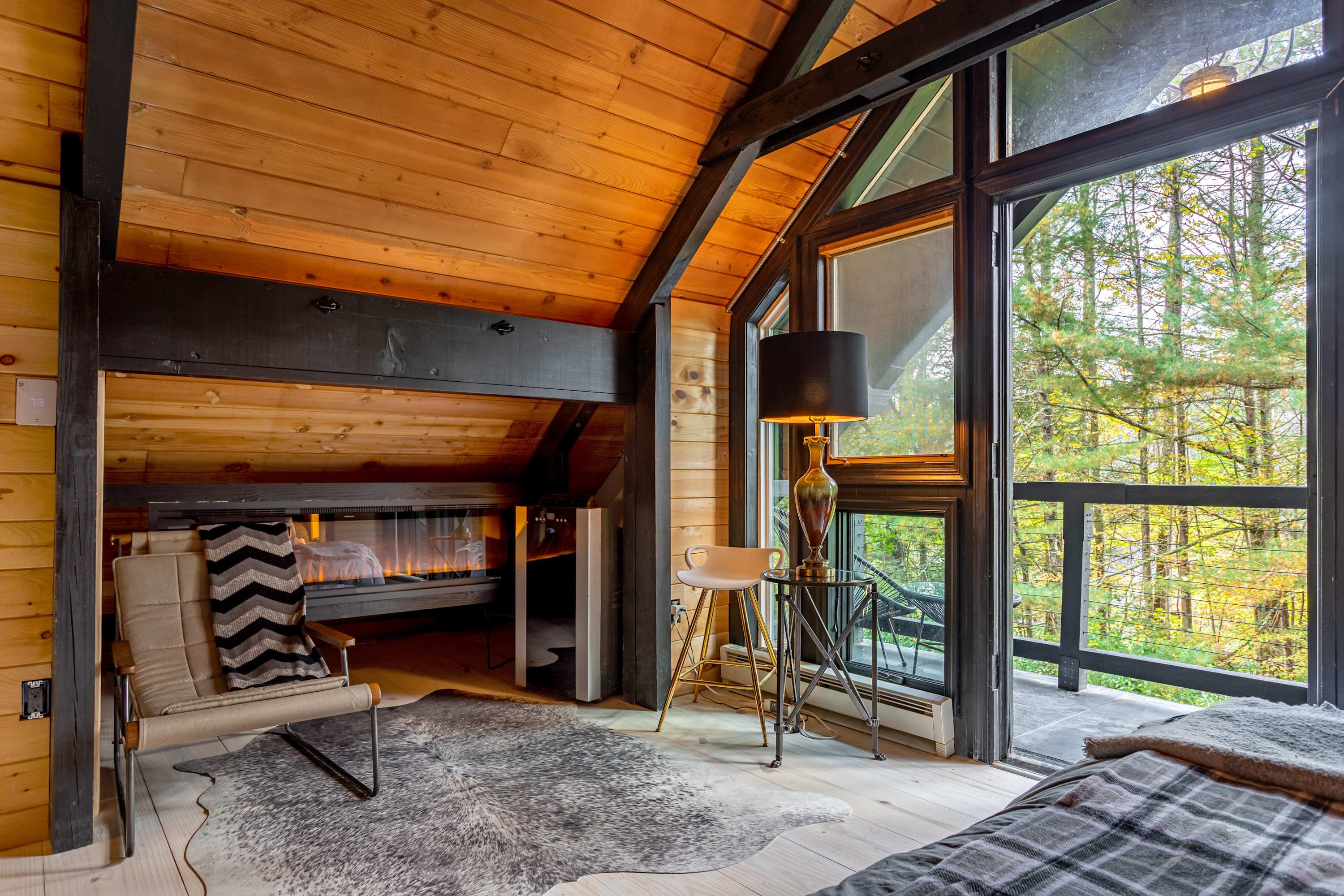 A cozy corner in a bedroom with a sloped wooden ceiling, featuring a modern armchair, a small side table with a lamp, and a linear fireplace built into the wooden wall. Large windows overlook the outdoors.
