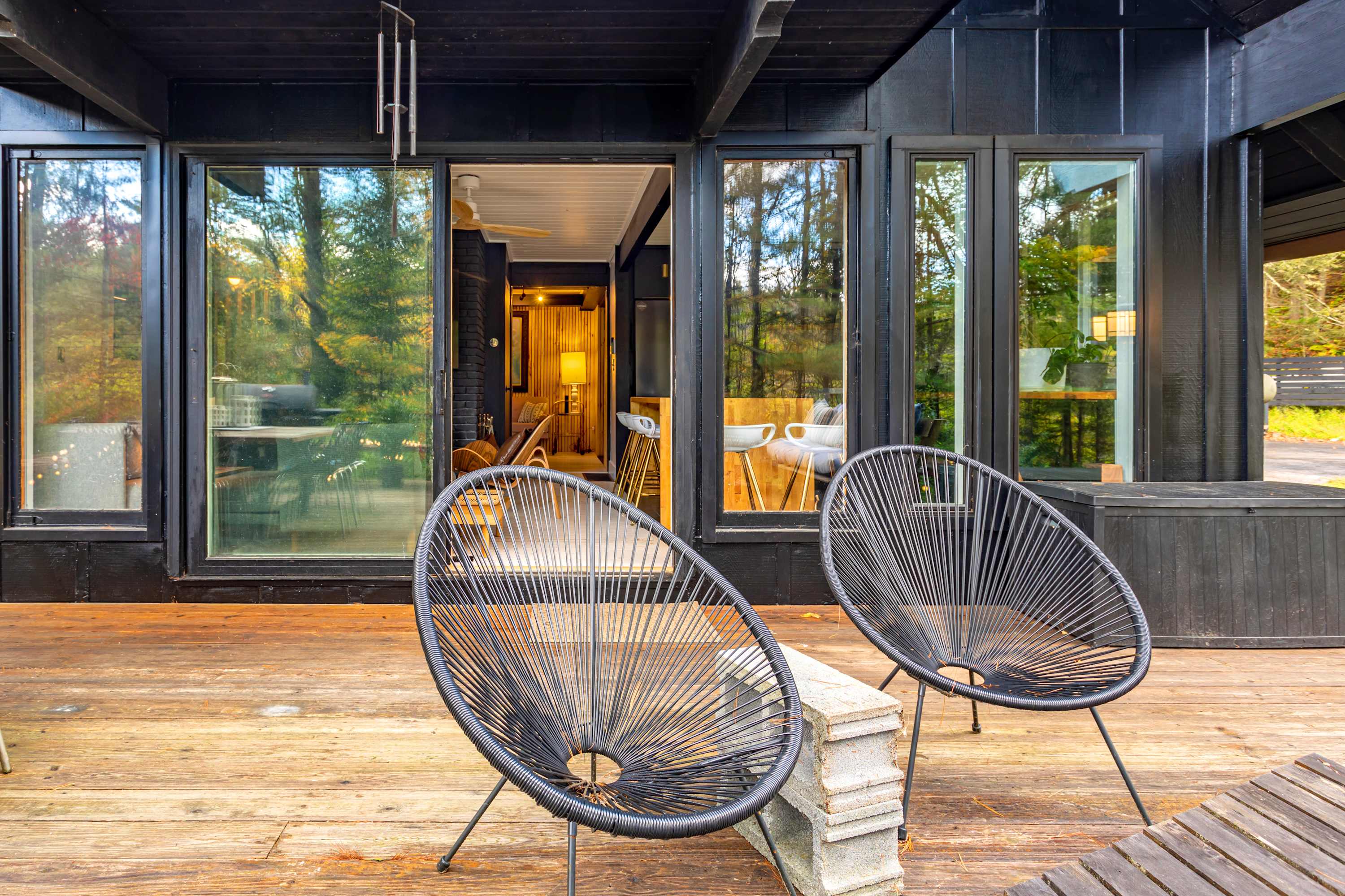 Wooden deck with two black wire chairs facing large glass sliding doors that lead into the house, offering a glimpse of an interior with a bar area. The house exterior is dark, blending with the surrounding trees.