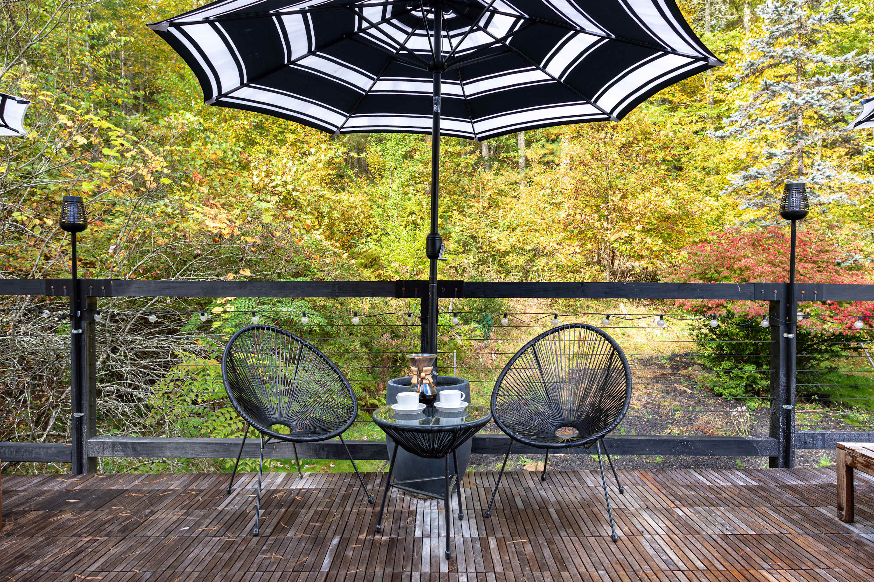 Cozy outdoor seating area on a wooden deck with two black wire chairs and a small round table, shaded by a large black and white striped umbrella. The area is surrounded by lush autumn trees and string lights.
