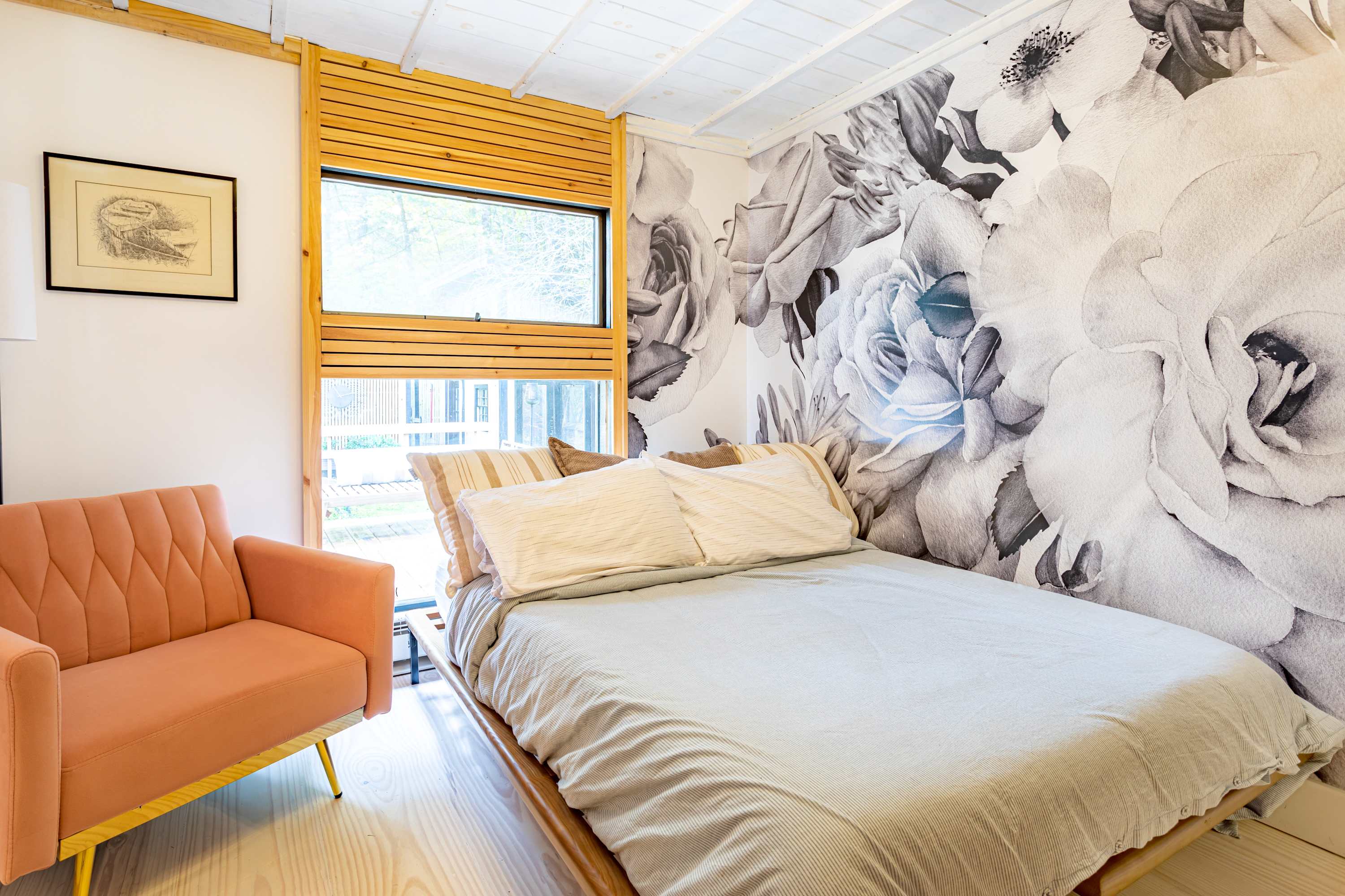 This bedroom features a large black and white floral mural wallpaper, a bed with light-colored bedding, and a peach-colored armchair with gold legs by a window with slatted wood blinds.