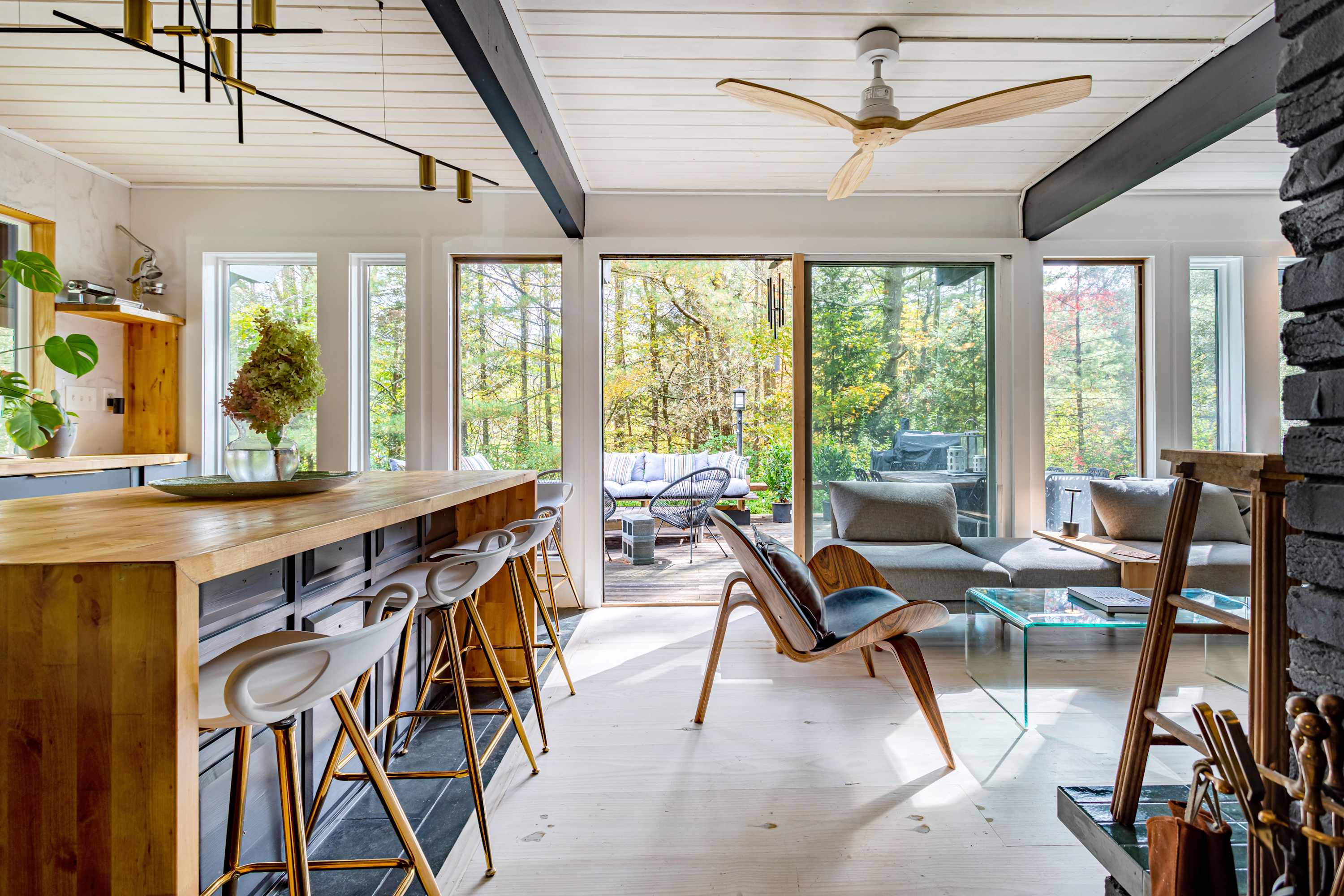 View from the kitchen island towards the living area and outdoor deck, showcasing the open layout with light hardwood floors, a wooden ceiling fan, and large windows offering forest views.