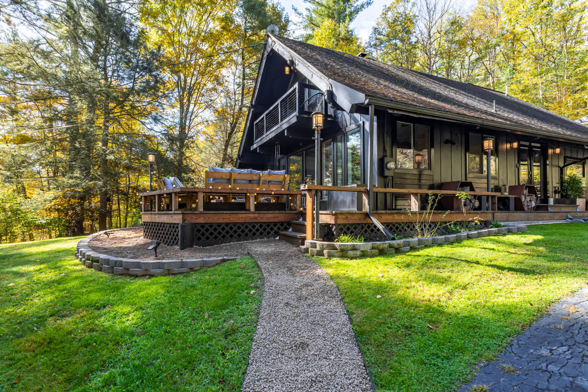 Dark-colored A-frame style house with a large wooden deck, surrounded by lush green grass and autumn trees with yellow foliage under a bright sky. A gravel path leads to the deck entrance.