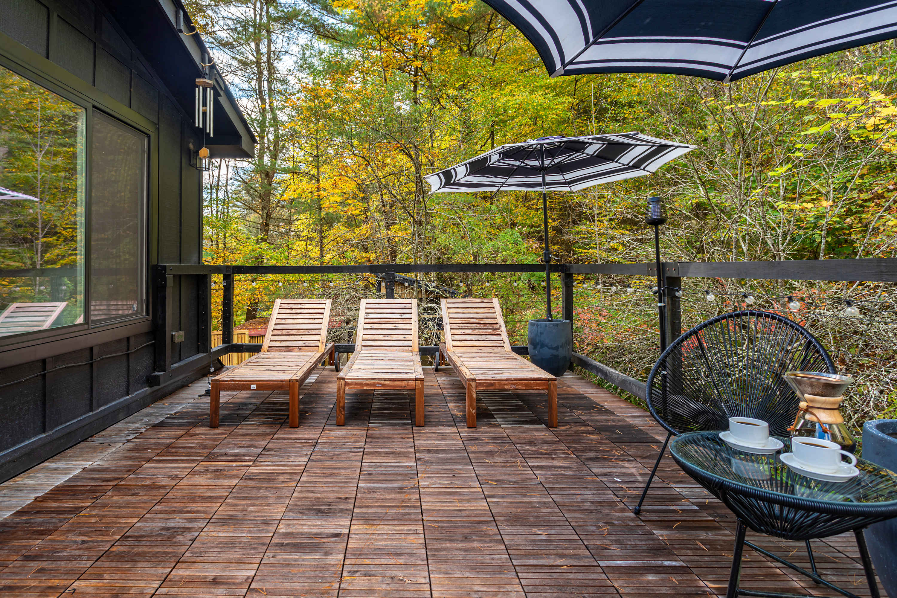 Expansive wooden deck featuring three wooden lounge chairs and a small black table with two black wire chairs under a striped umbrella. The deck overlooks a vibrant autumn forest.