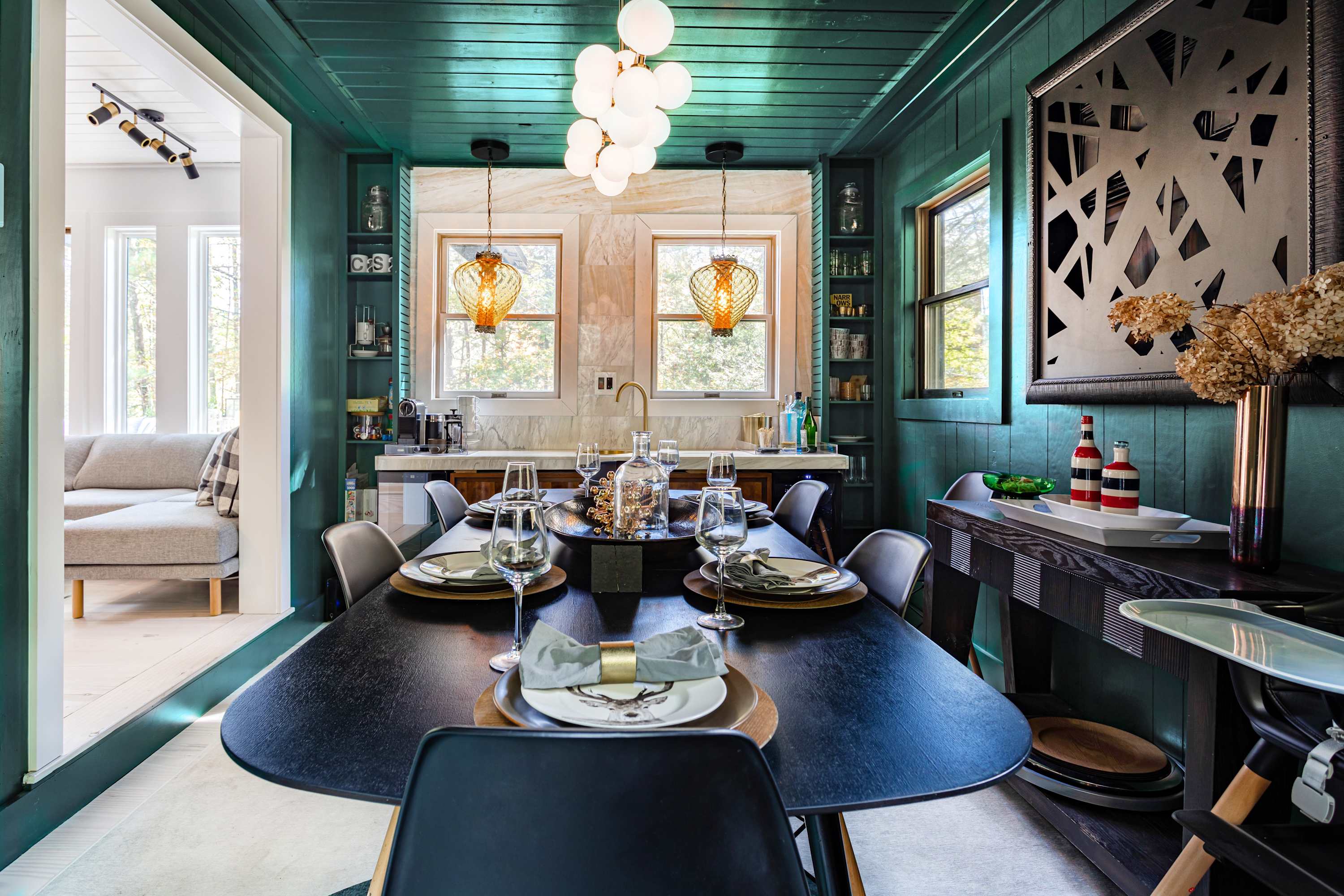 Elegant dining room with dark green walls and ceiling, a large black oval dining table with modern chairs, and a unique globe chandelier, with a view into the kitchen.