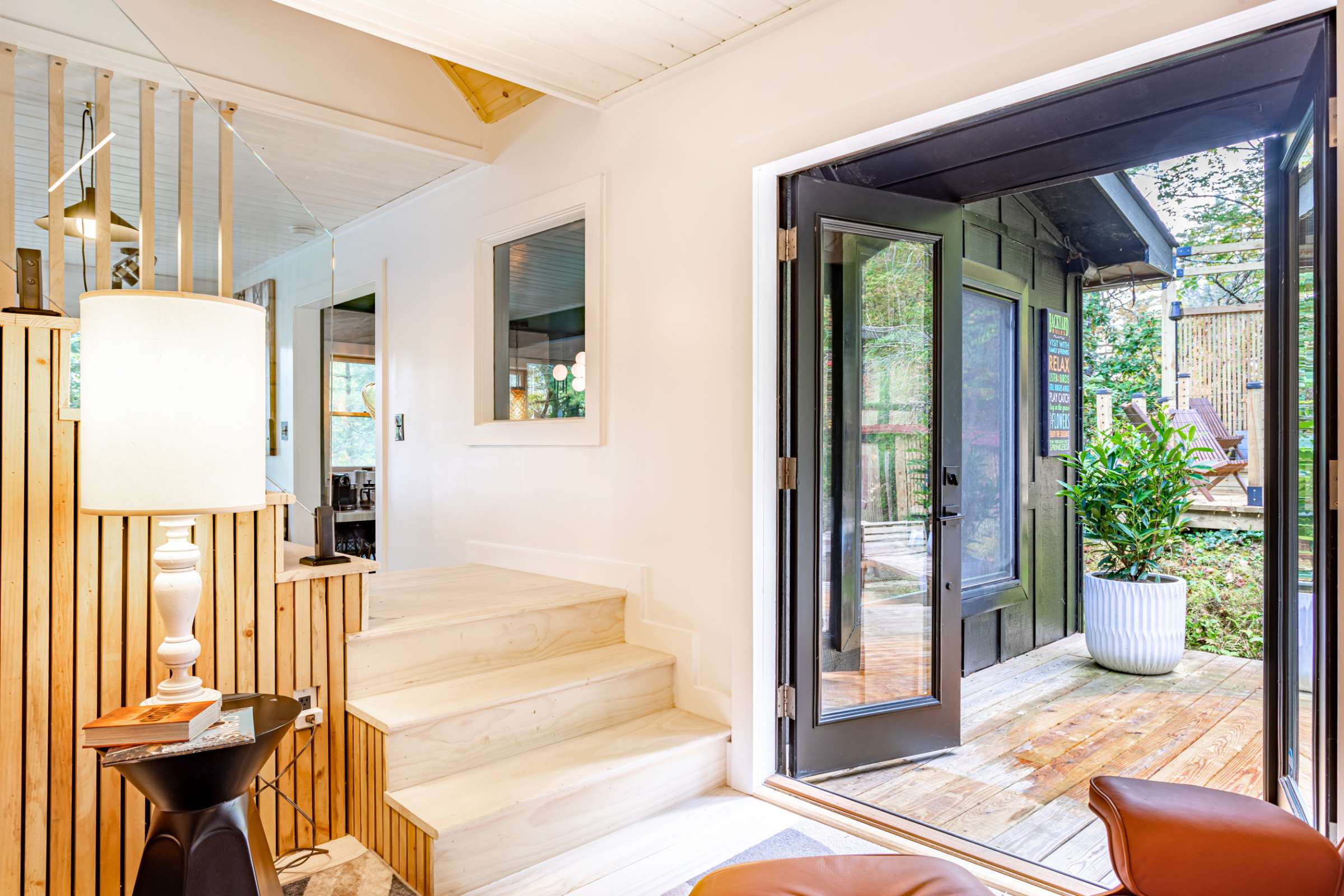 Entryway with light wooden stairs and a slatted railing, leading to an upper level, with an open doorway revealing an outdoor deck and lush greenery.