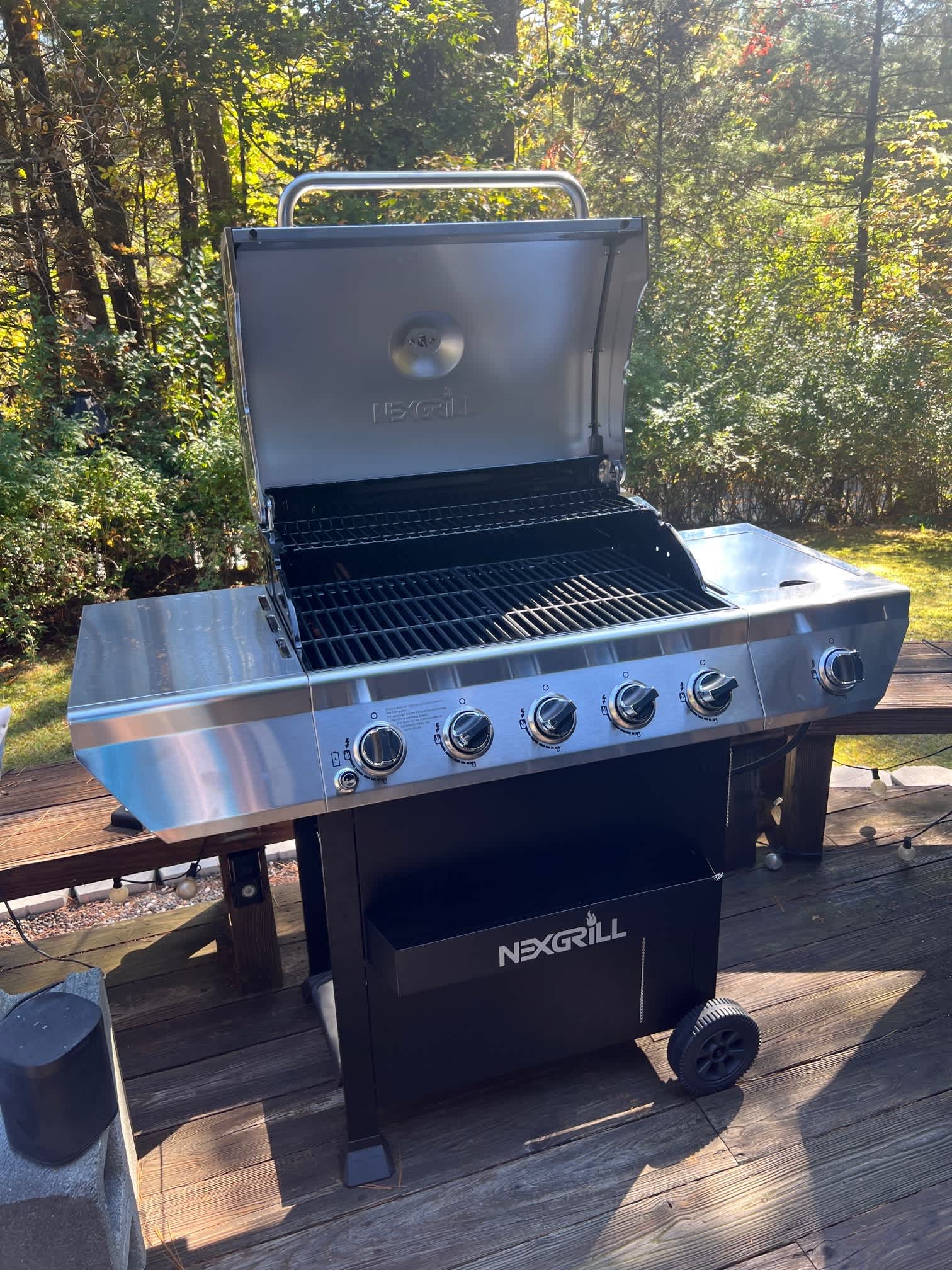 A modern stainless steel and black gas grill, branded 'NEXGRILL', stands with its lid open on a rustic wooden deck, revealing the cooking grates and control panel. Lush green trees and foliage provide a natural backdrop to this outdoor cooking area.