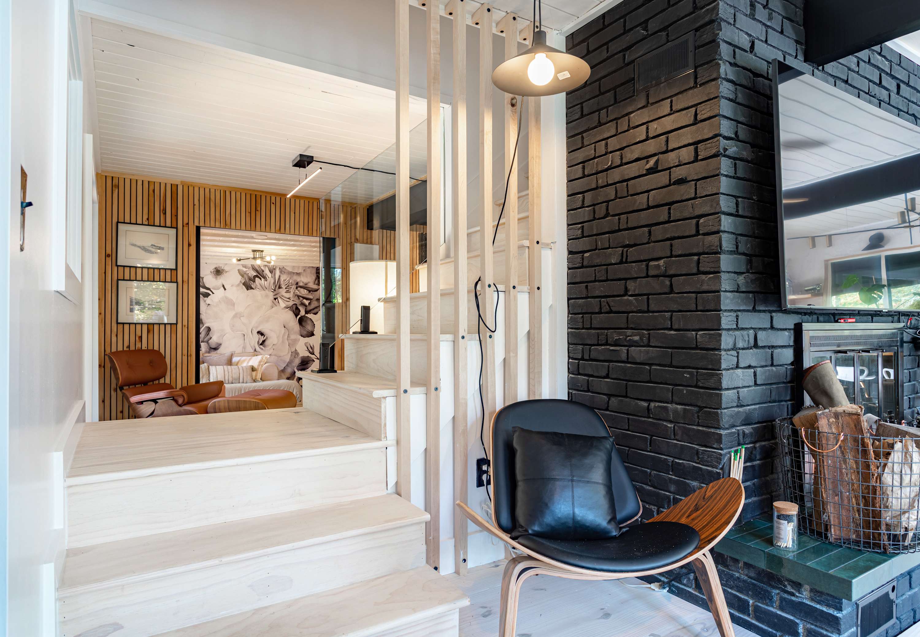 Bright entryway with light wooden stairs and a slatted wooden railing, leading to an upper level, featuring a black brick fireplace and a modern wooden chair with a black cushion.
