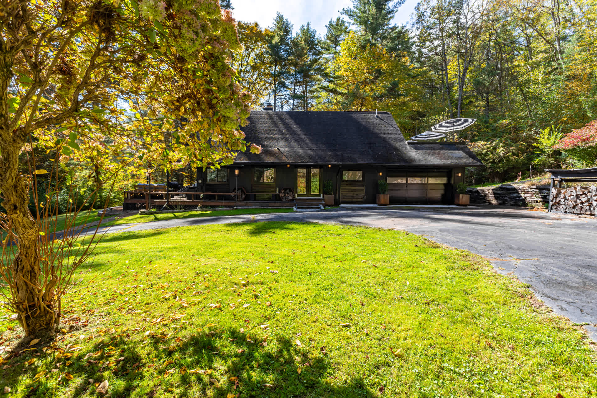 Front view of the dark-colored chalet-style house with a large driveway and a well-maintained green lawn, surrounded by mature trees displaying beautiful autumn foliage. Striped umbrellas are visible on an upper deck.