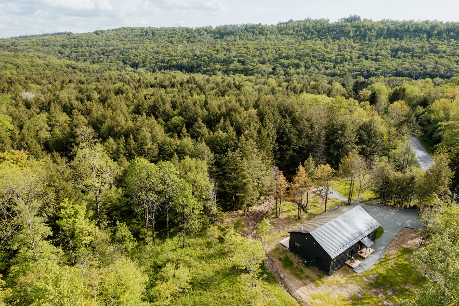 Aerial view of a dark gray modern cabin with a metal roof, situated amidst a lush green forest, with a gravel driveway leading to the property.