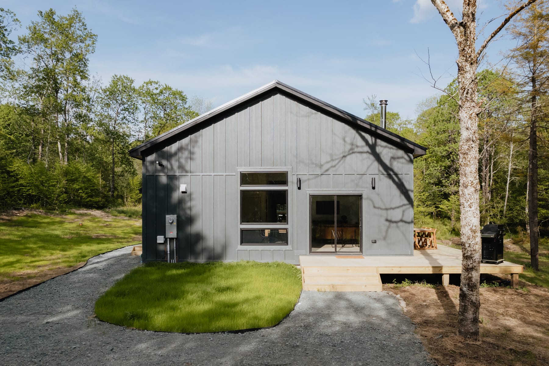 The front facade of a modern dark gray cabin with vertical siding, featuring a large sliding glass door and a multi-pane window, with a small wooden deck and steps leading to a gravel path.
