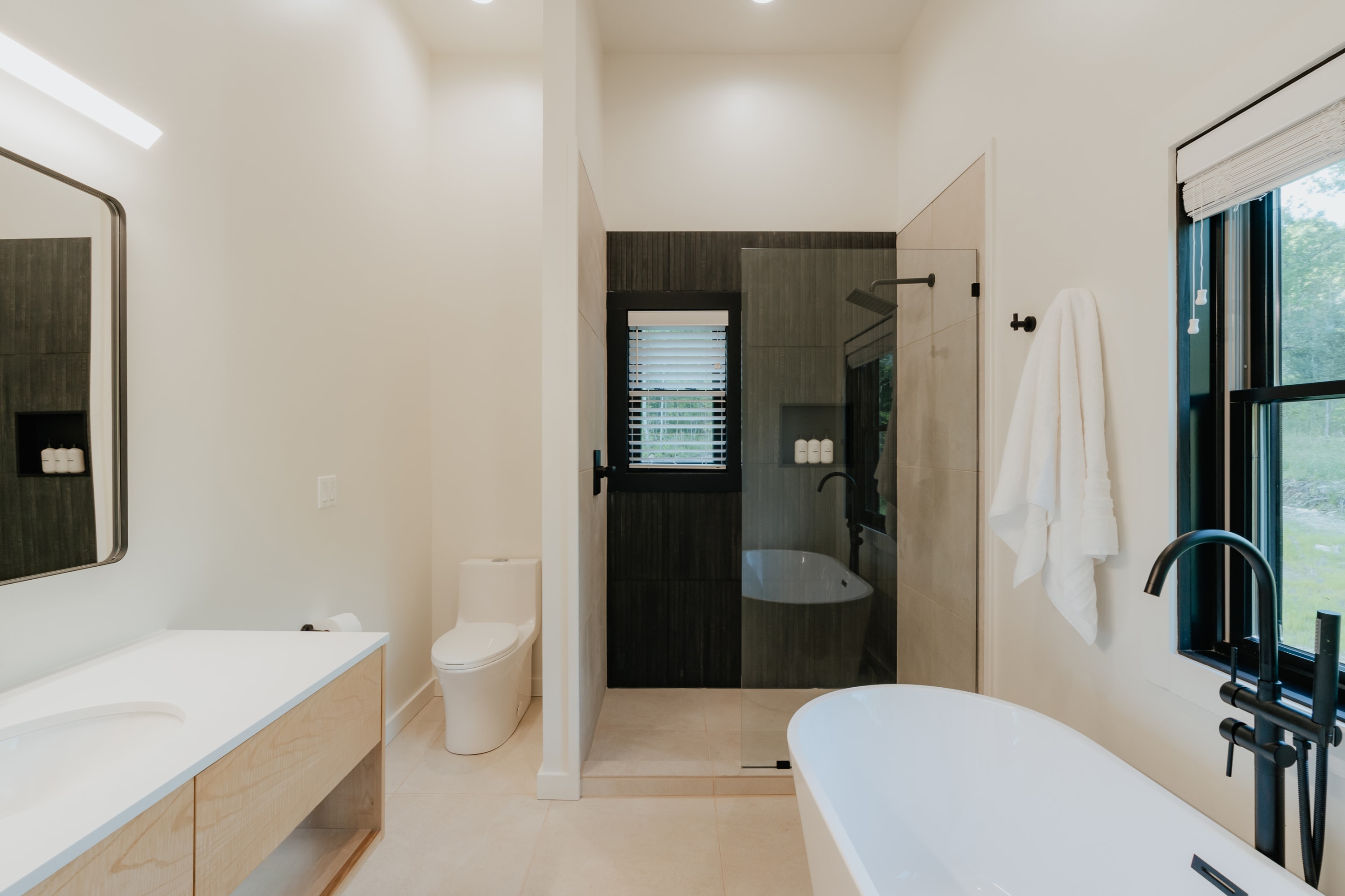 Modern bathroom featuring a light wood vanity with a white countertop and rectangular mirror, a toilet, a glass-enclosed shower with dark tiled walls, and a white freestanding bathtub with a black floor-mounted faucet.