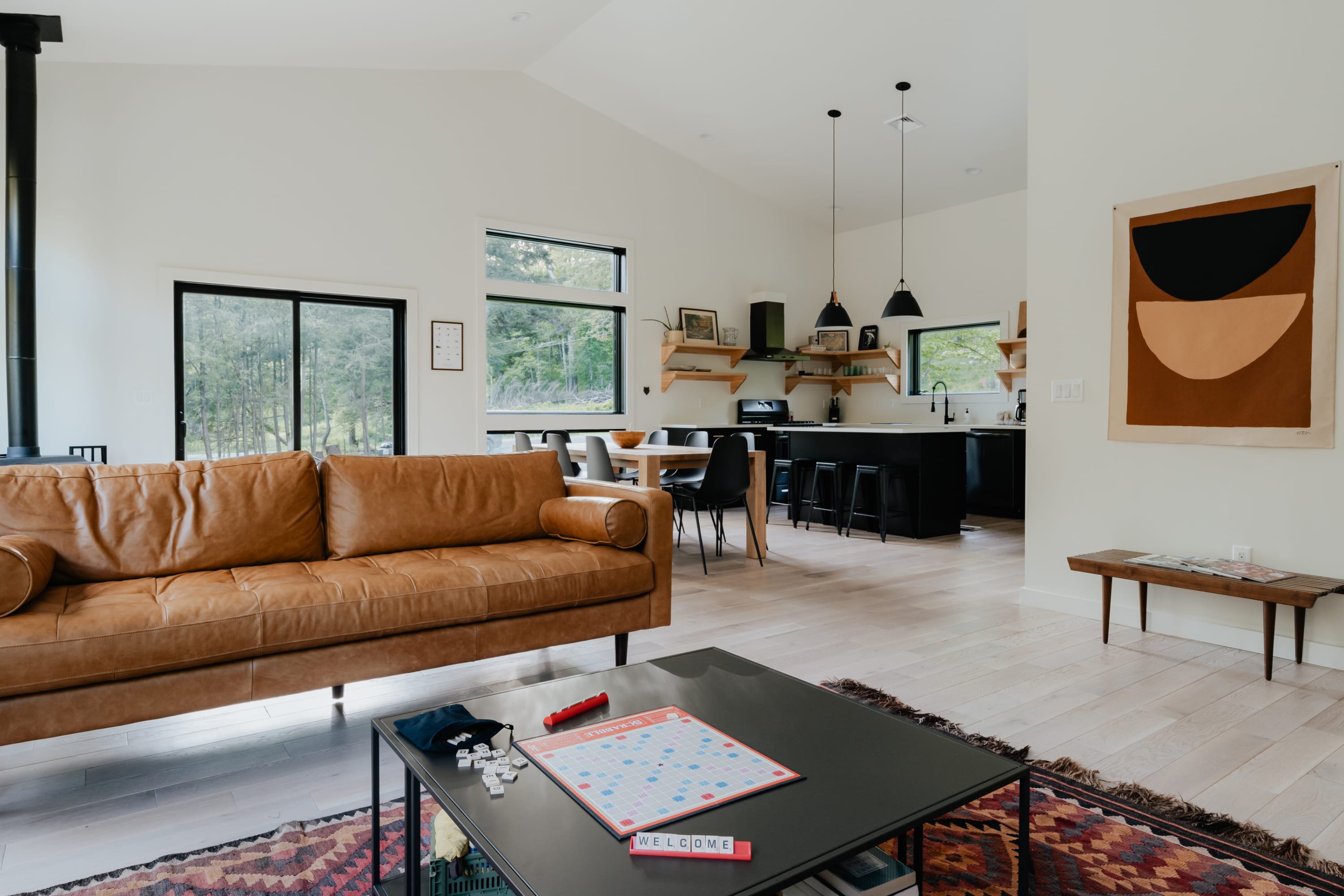 Cozy living room with a large brown leather sofa, a black coffee table with a board game, and a colorful rug. The open layout connects to the dining and kitchen areas, with large windows offering views of the surrounding nature.