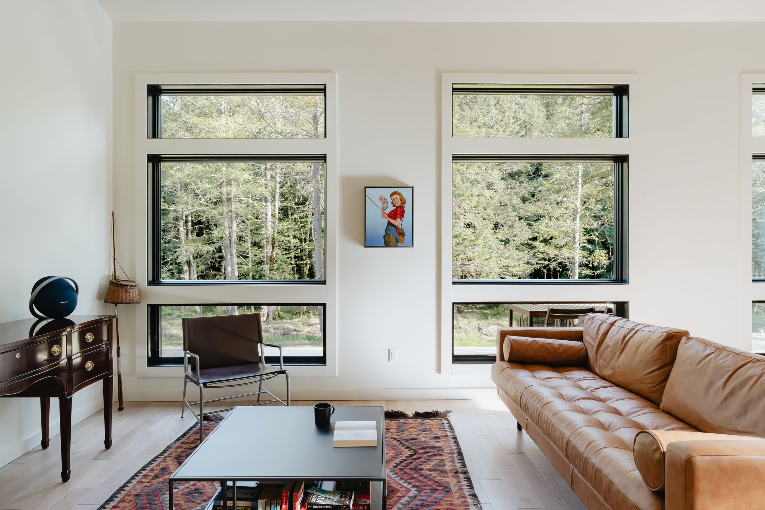 Bright living room featuring a tan leather sofa, a black coffee table on a patterned rug, and multiple large windows offering views of a lush forest, with a unique painting on the wall.