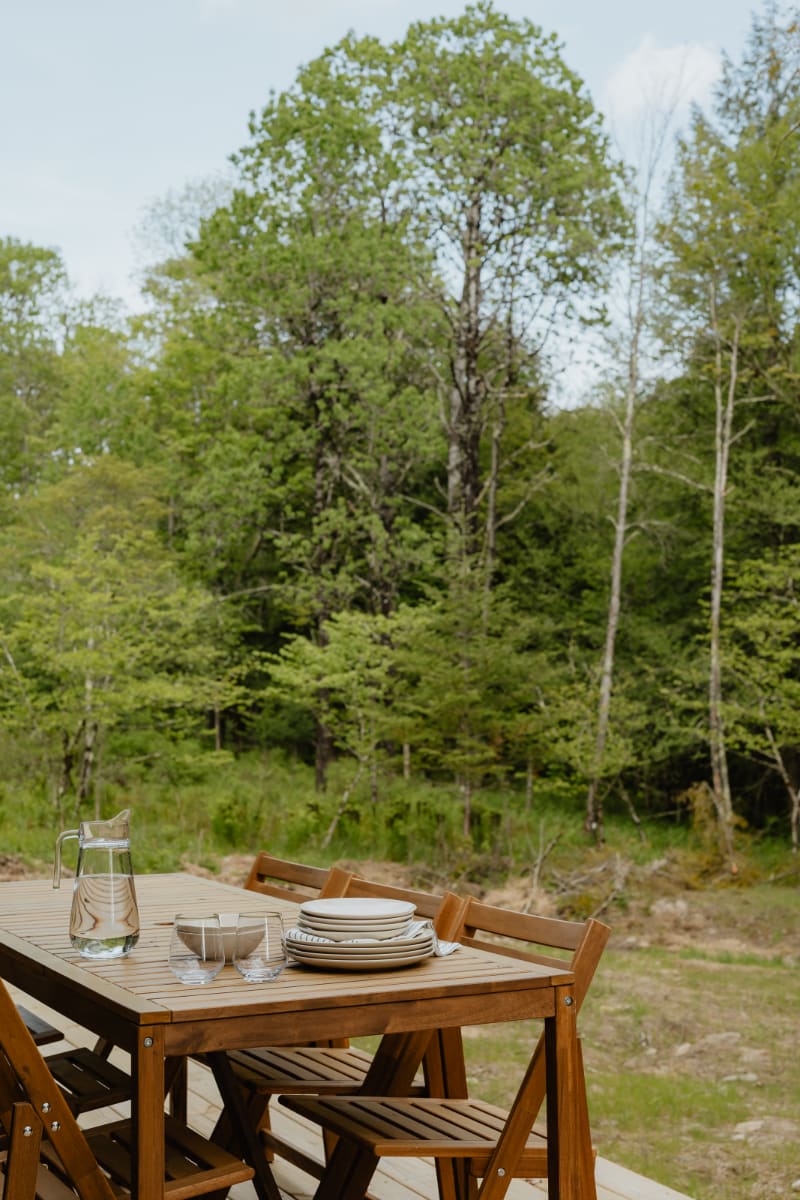 A wooden outdoor dining table with chairs is set on a deck, ready for a meal, with a pitcher of water and plates. Lush green trees and foliage create a serene natural backdrop.