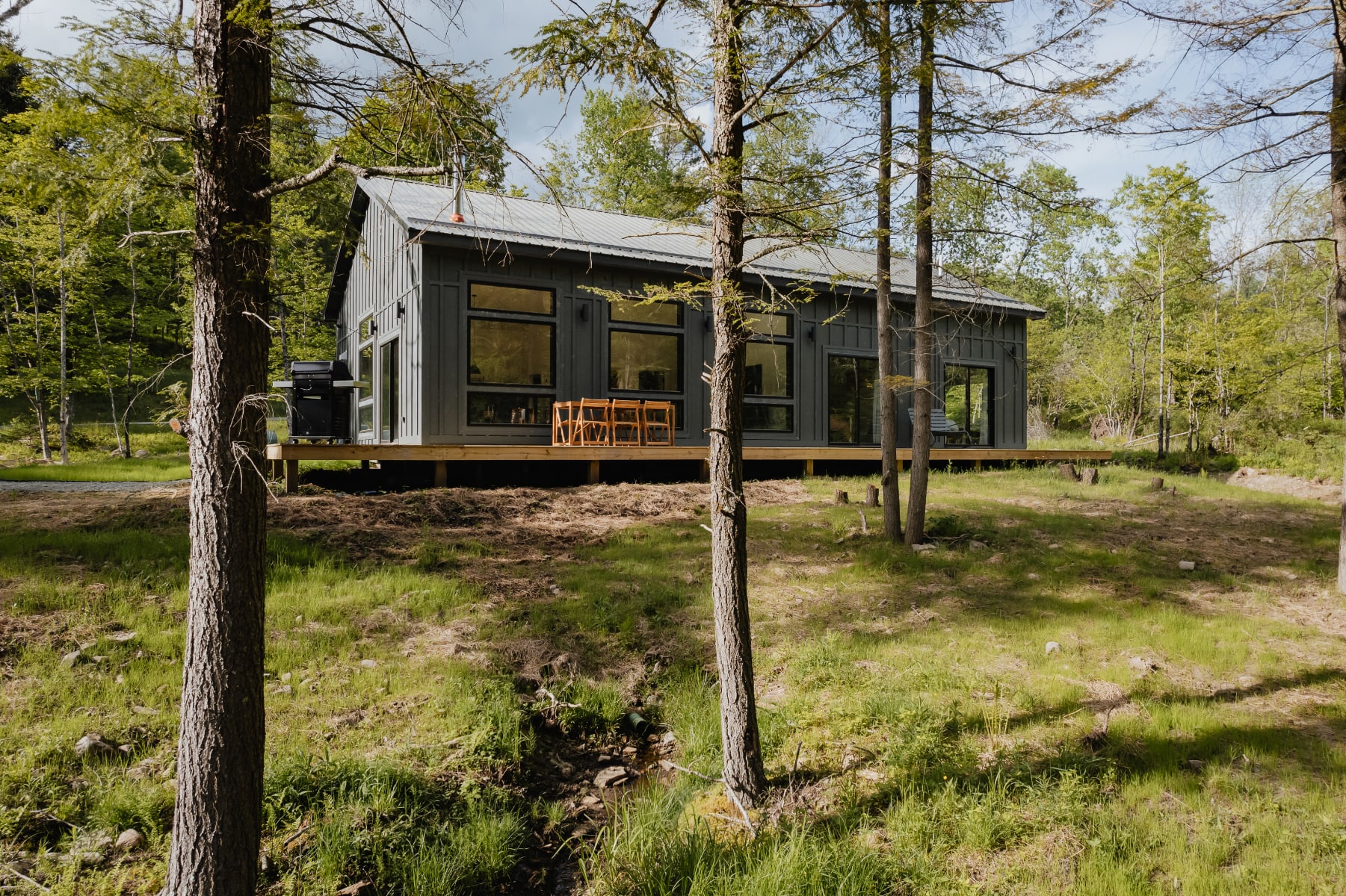 The back of a modern dark gray cabin with extensive windows, opening onto a spacious wooden deck furnished with outdoor seating and a BBQ grill, nestled among tall trees and green grass.