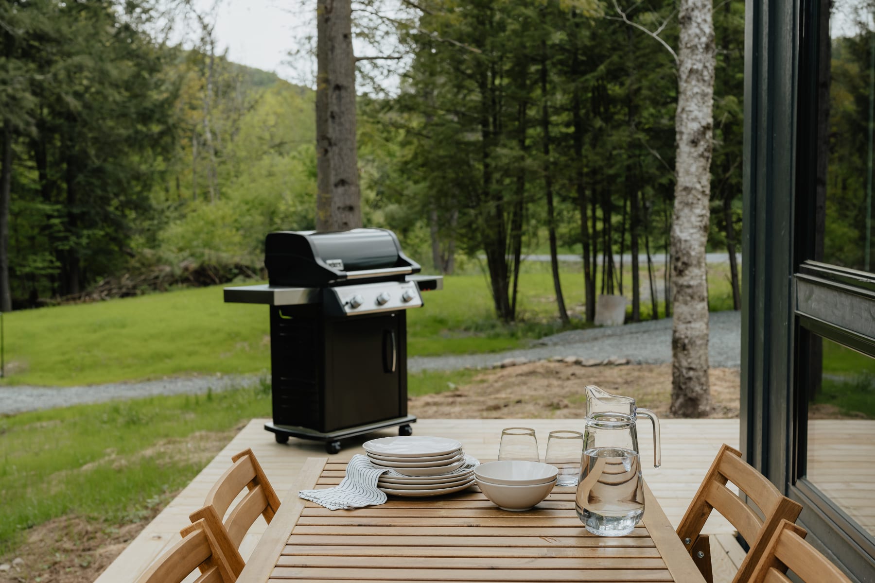 Wooden outdoor deck with a dining table set with plates, bowls, and a pitcher, accompanied by wooden chairs, and a black gas grill visible in the background against a lush green lawn and trees.