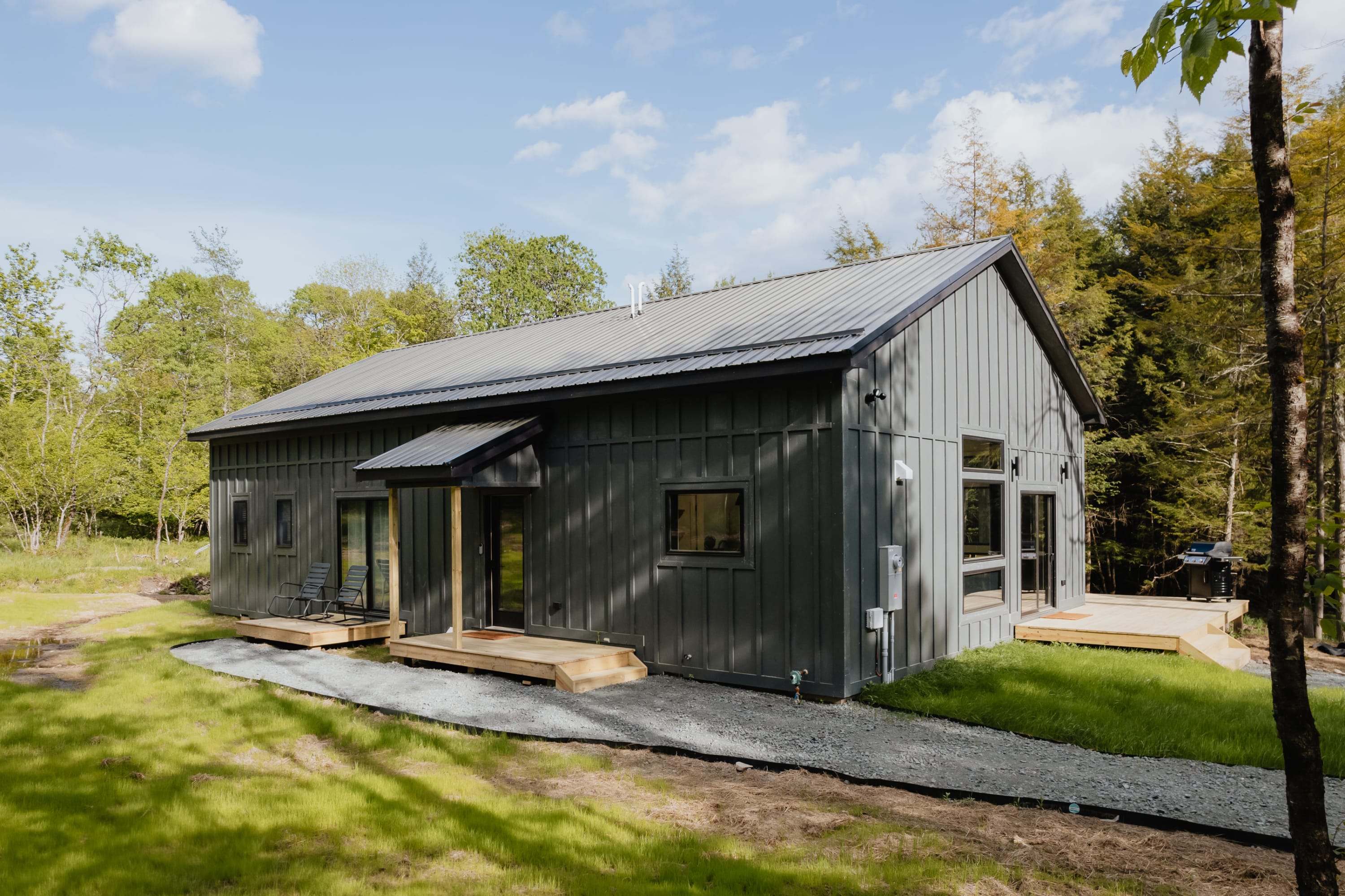 Modern dark gray cabin with vertical siding and a metal roof, featuring multiple large windows and a small covered porch entrance, surrounded by green grass and trees.