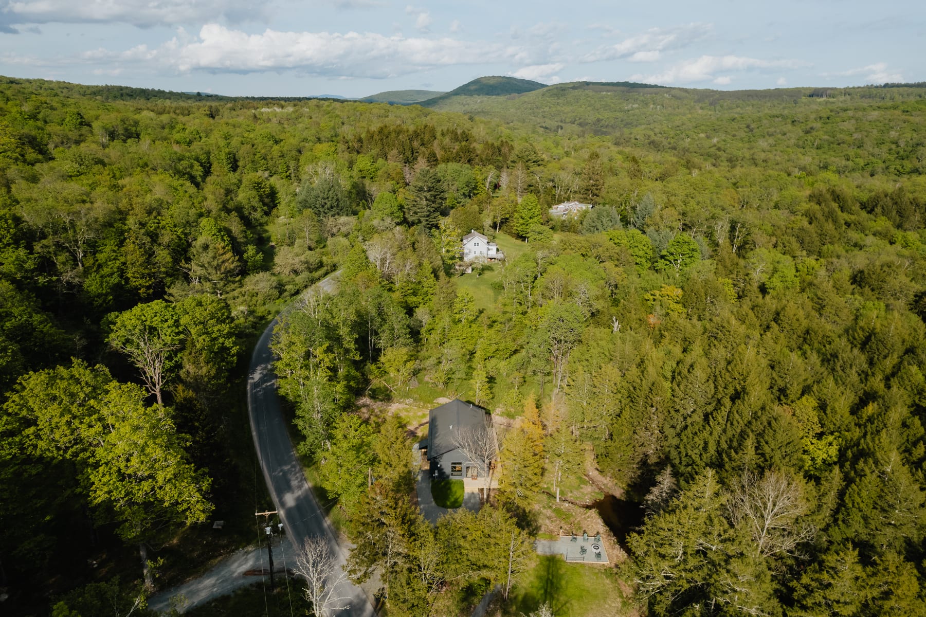 Wide aerial view showcasing a modern dark gray cabin nestled within a vast, verdant forest landscape with rolling hills and a winding road, highlighting its secluded natural setting.