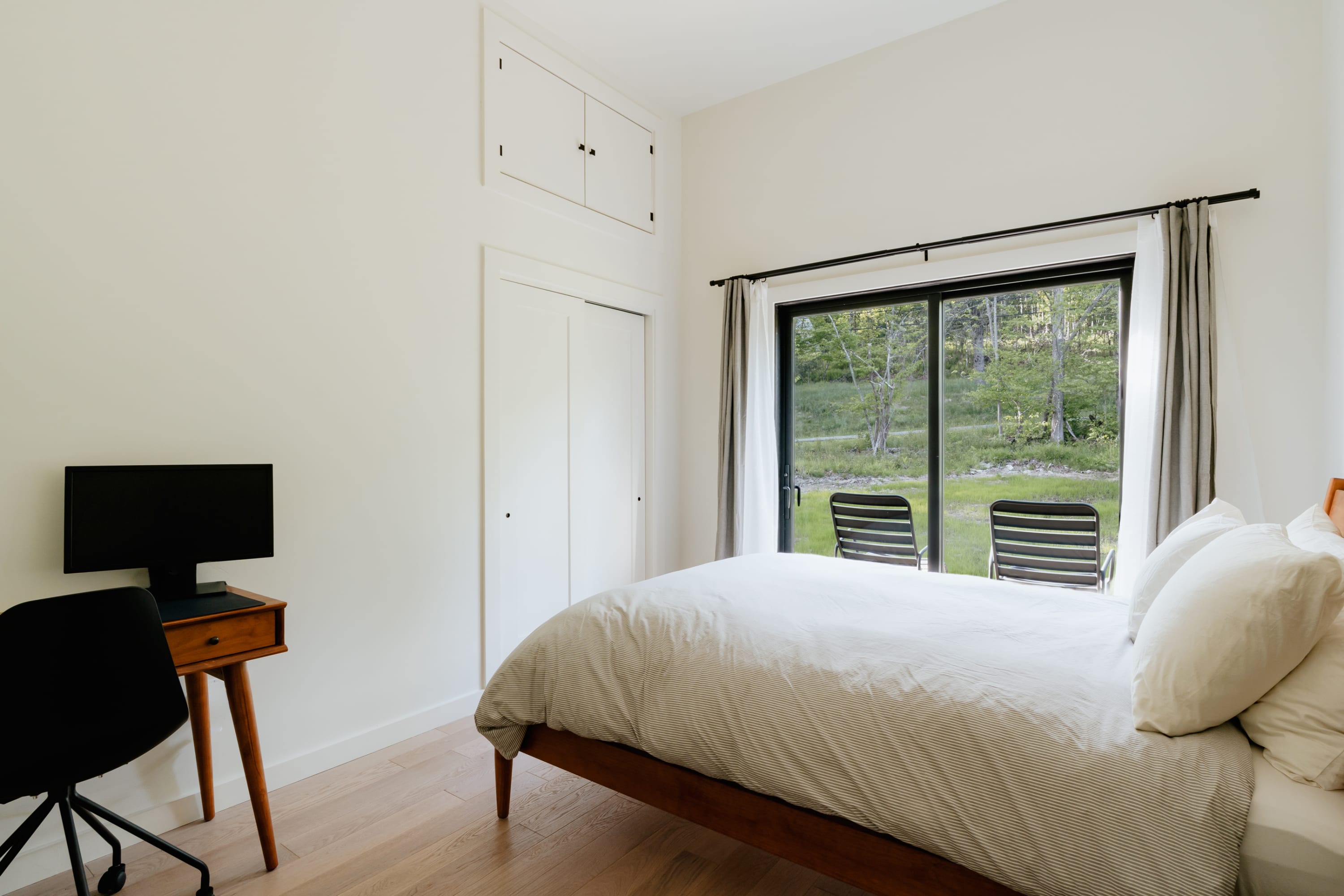 Bright bedroom with a wooden bed frame, white and grey striped duvet, a small wooden desk with a black office chair and monitor, and large sliding glass doors opening to a grassy backyard with two lounge chairs and trees.