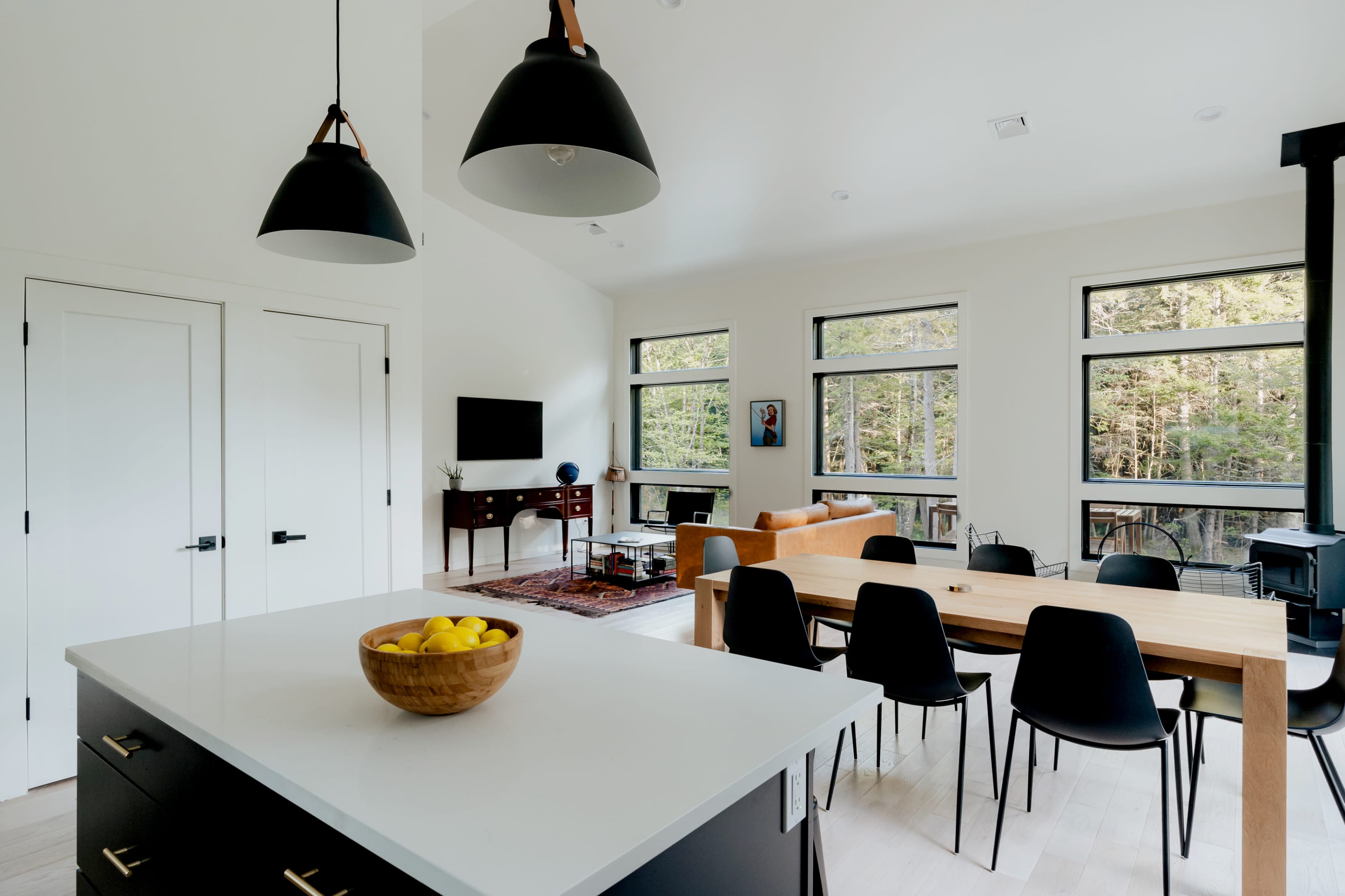 Open-concept living and dining area with a light wood dining table and black chairs under pendant lights, adjacent to a kitchen island and a living room visible in the background, all with forest views.