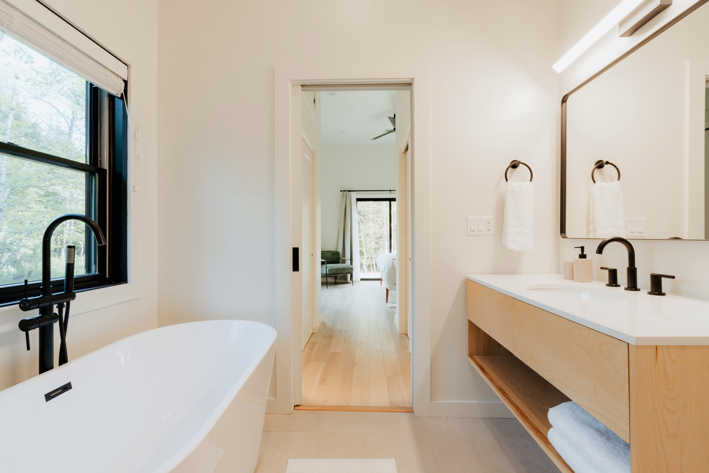 Modern bathroom with a freestanding white bathtub, a wooden vanity with a white sink and black faucet, and a large mirror, with a view into a bedroom through an open doorway.