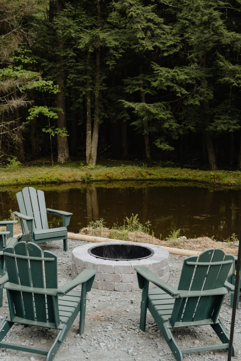 Outdoor fire pit area with a circular stone fire ring surrounded by several dark green Adirondack chairs, situated next to a calm pond and backed by a dense forest of evergreen trees.