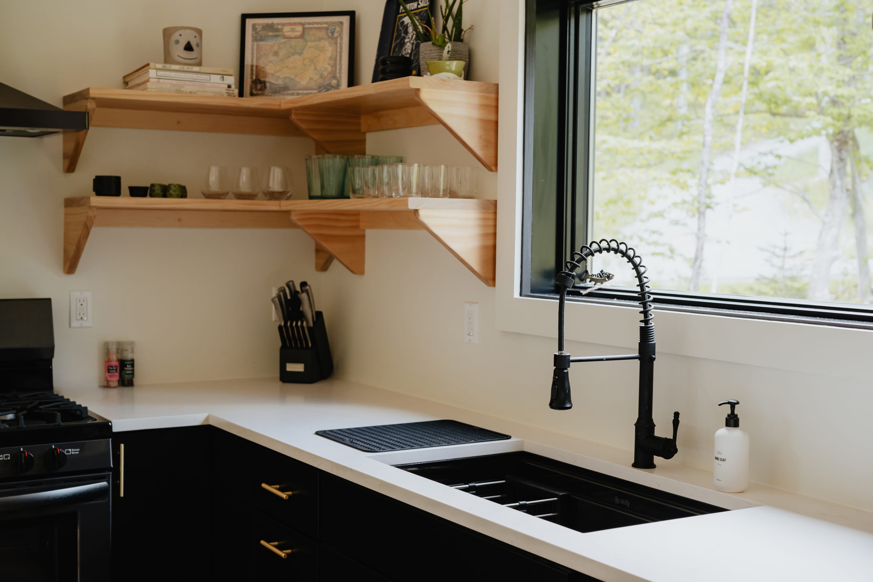 Close-up of a modern kitchen corner with black cabinets, white countertops, and a black gooseneck faucet over a black sink, complemented by open wooden shelves holding glassware and a knife block.