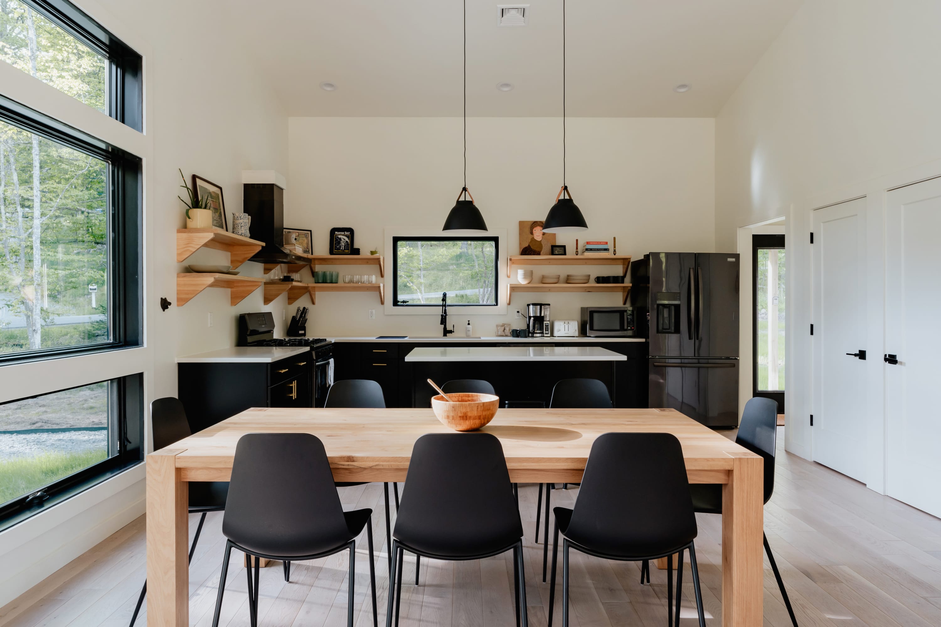 Wide view of the open-plan dining and kitchen area, featuring a light wood dining table with black chairs, a modern kitchen with black appliances and open shelving, and large windows.