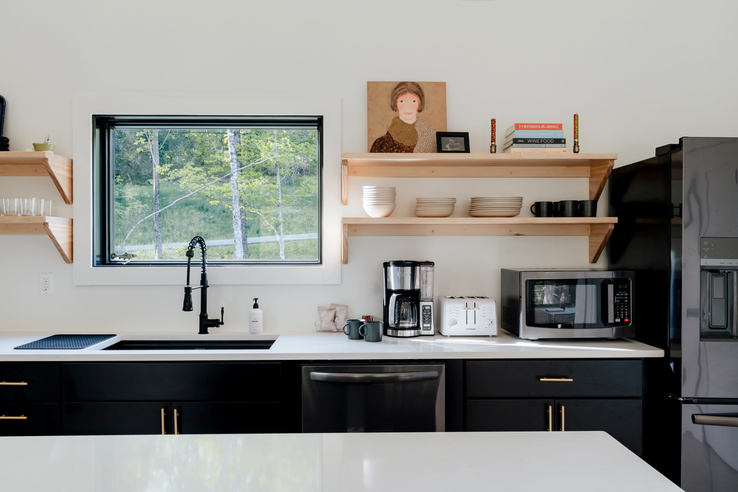 Well-lit kitchen area with black lower cabinets, white countertops, and open wooden shelving, featuring a sink under a large window with a forest view, and various small appliances like a coffee maker and microwave.