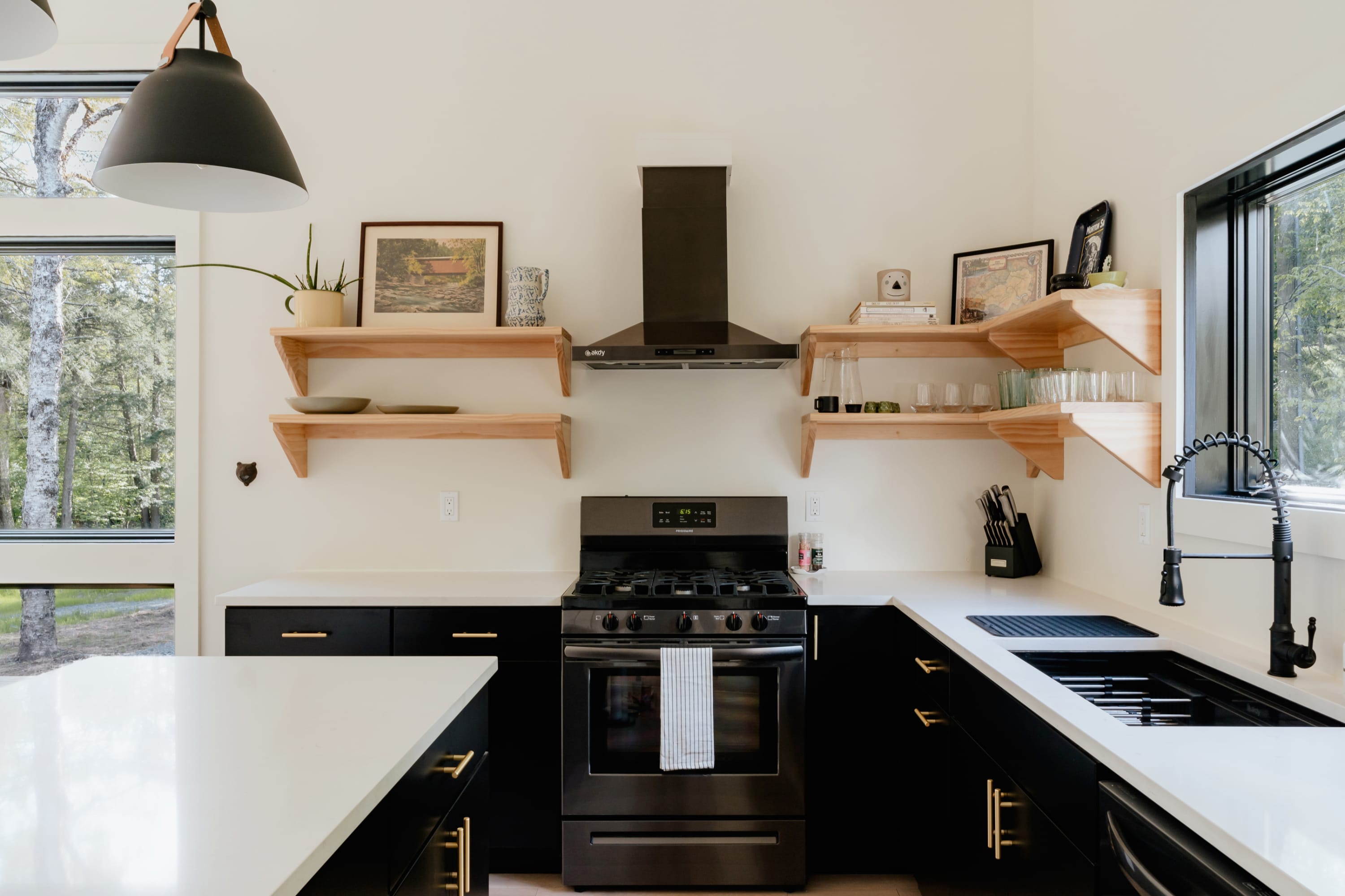 Modern kitchen with black cabinetry, white countertops, and a black gas range with a matching hood, flanked by open wooden shelves, all under a large pendant light.