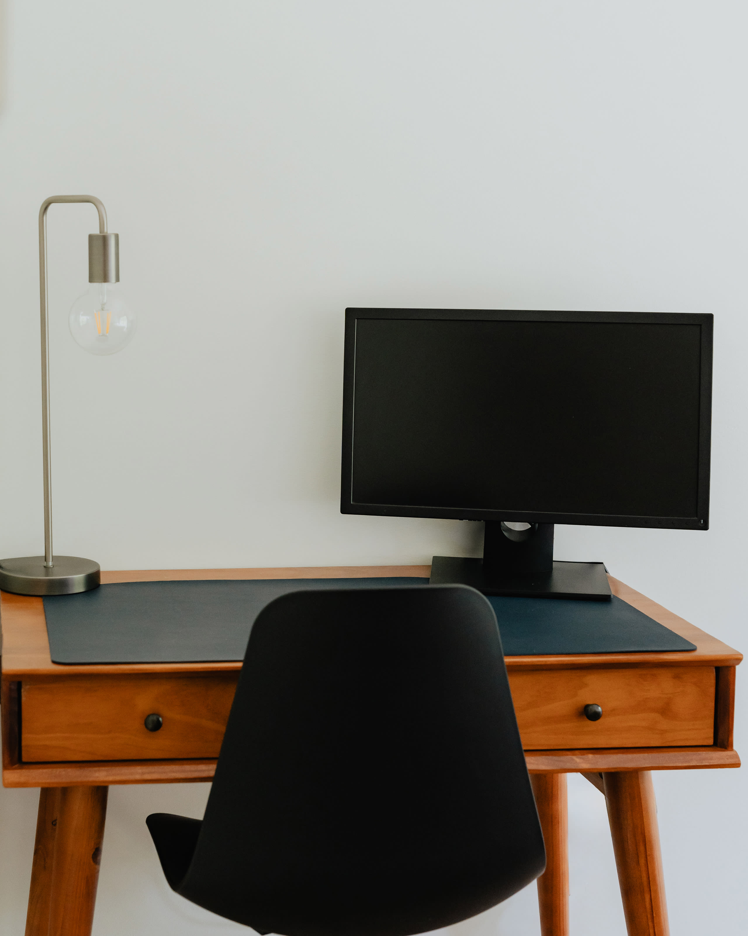 Modern wooden desk with a dark blue desk pad, a black office chair, a silver desk lamp with an exposed bulb, and a black computer monitor against a white wall.