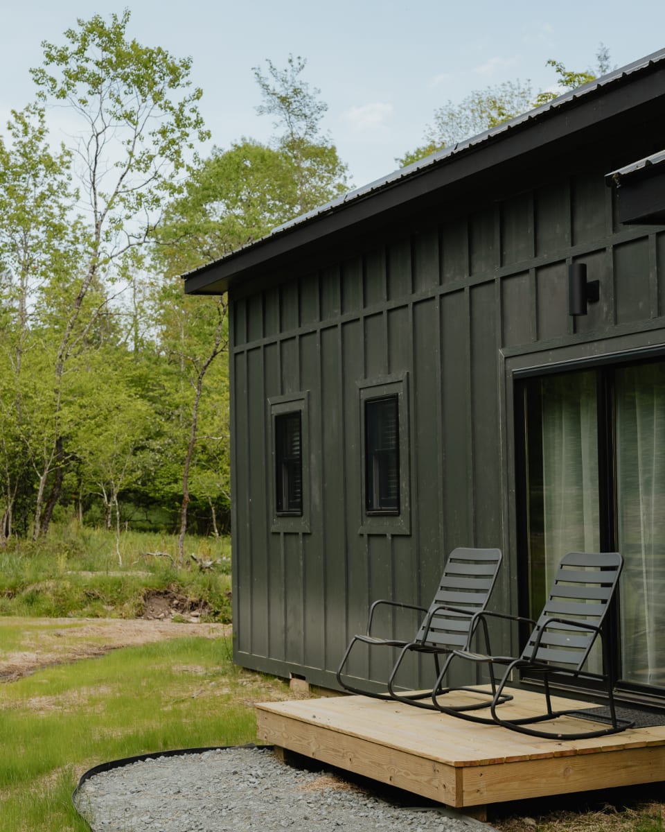 Side view of a modern dark green cabin with vertical siding and black-framed windows, featuring a small wooden deck with two dark grey rocking chairs, overlooking a grassy area and surrounding trees.