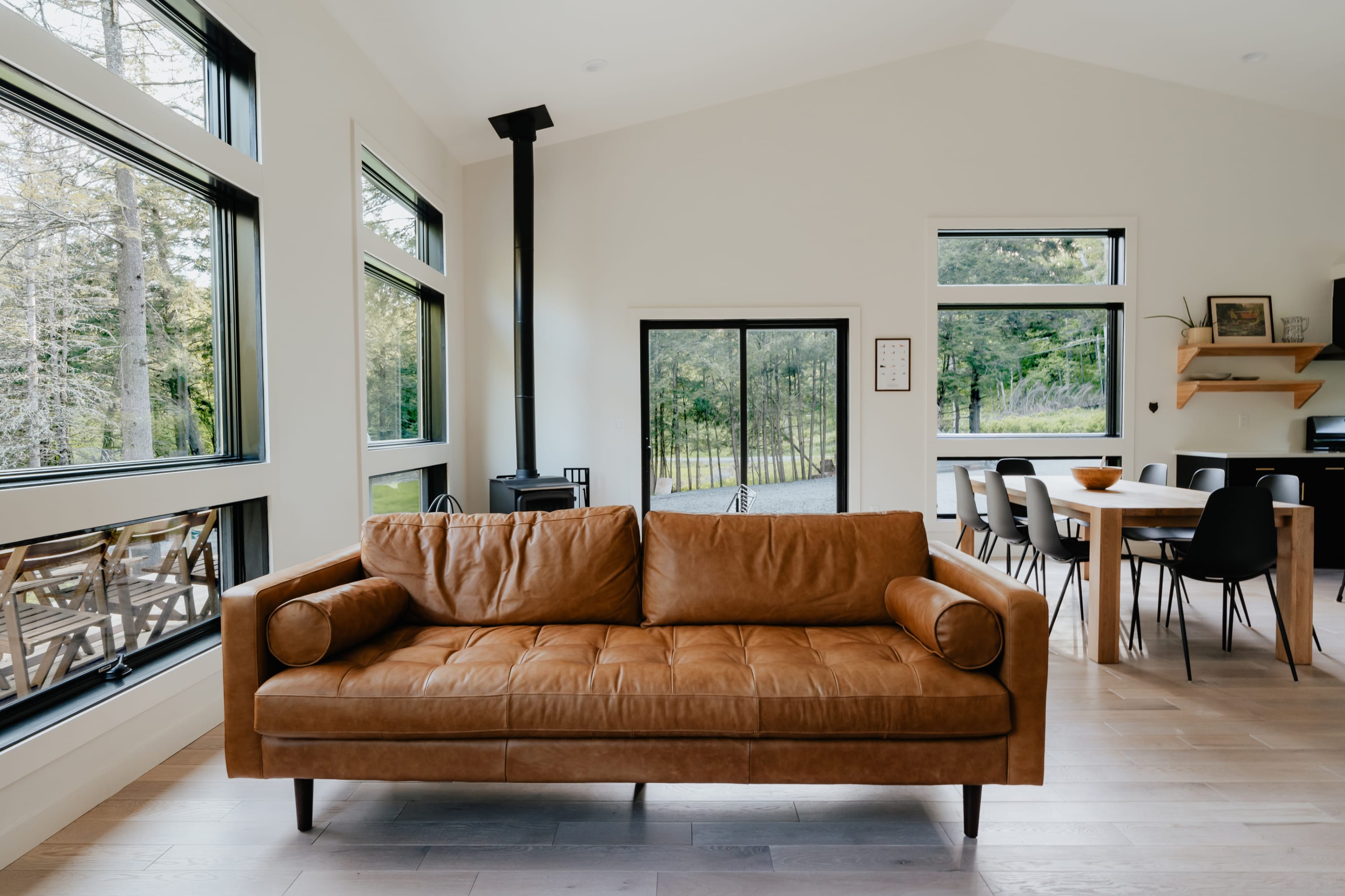 Spacious living room with a large tan leather sofa facing a wood-burning stove, light hardwood floors, and expansive windows showcasing the surrounding forest, leading into a dining area.