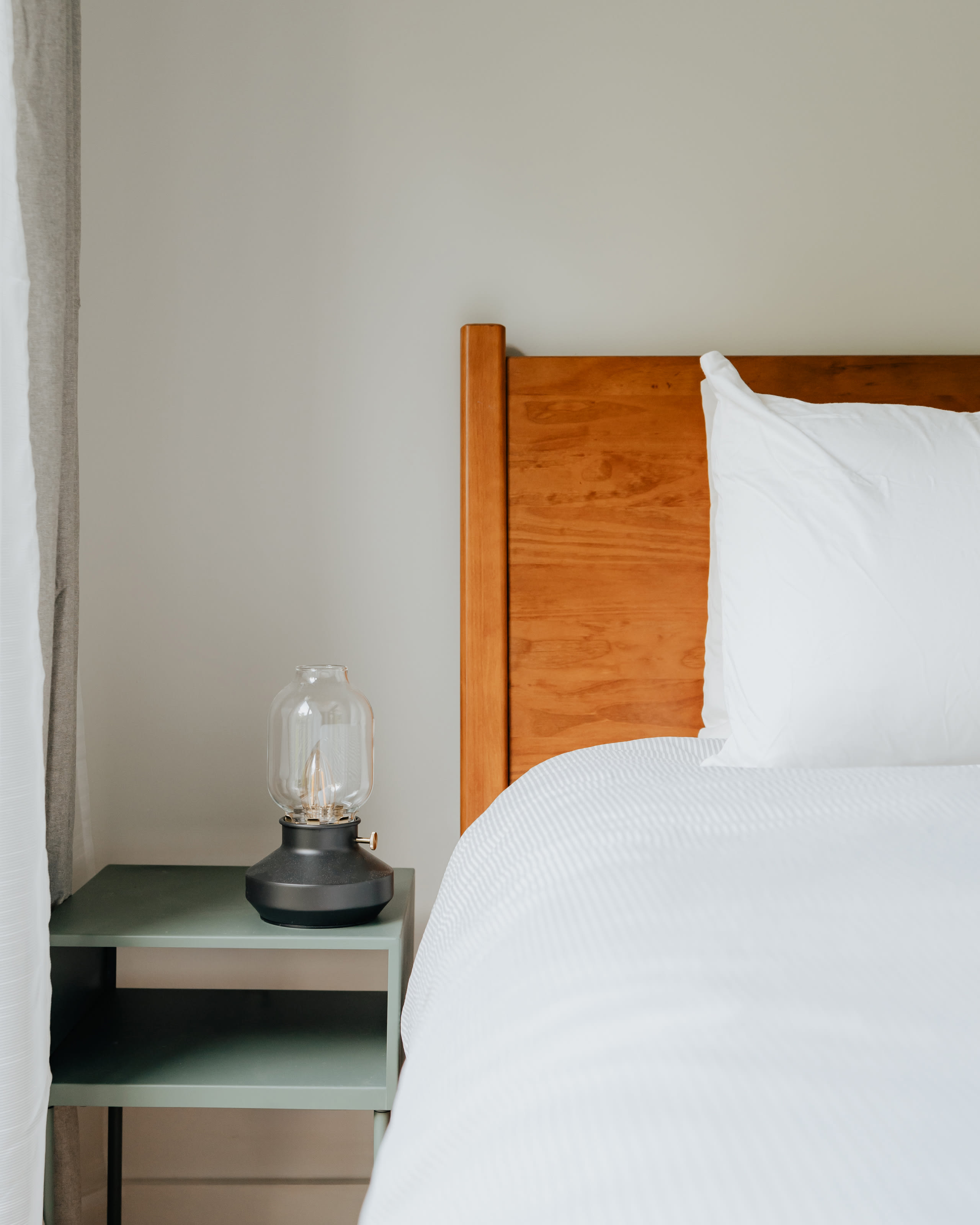 Close-up of a wooden headboard and a white bed with a textured duvet, next to a light green bedside table holding a modern lantern-style lamp.